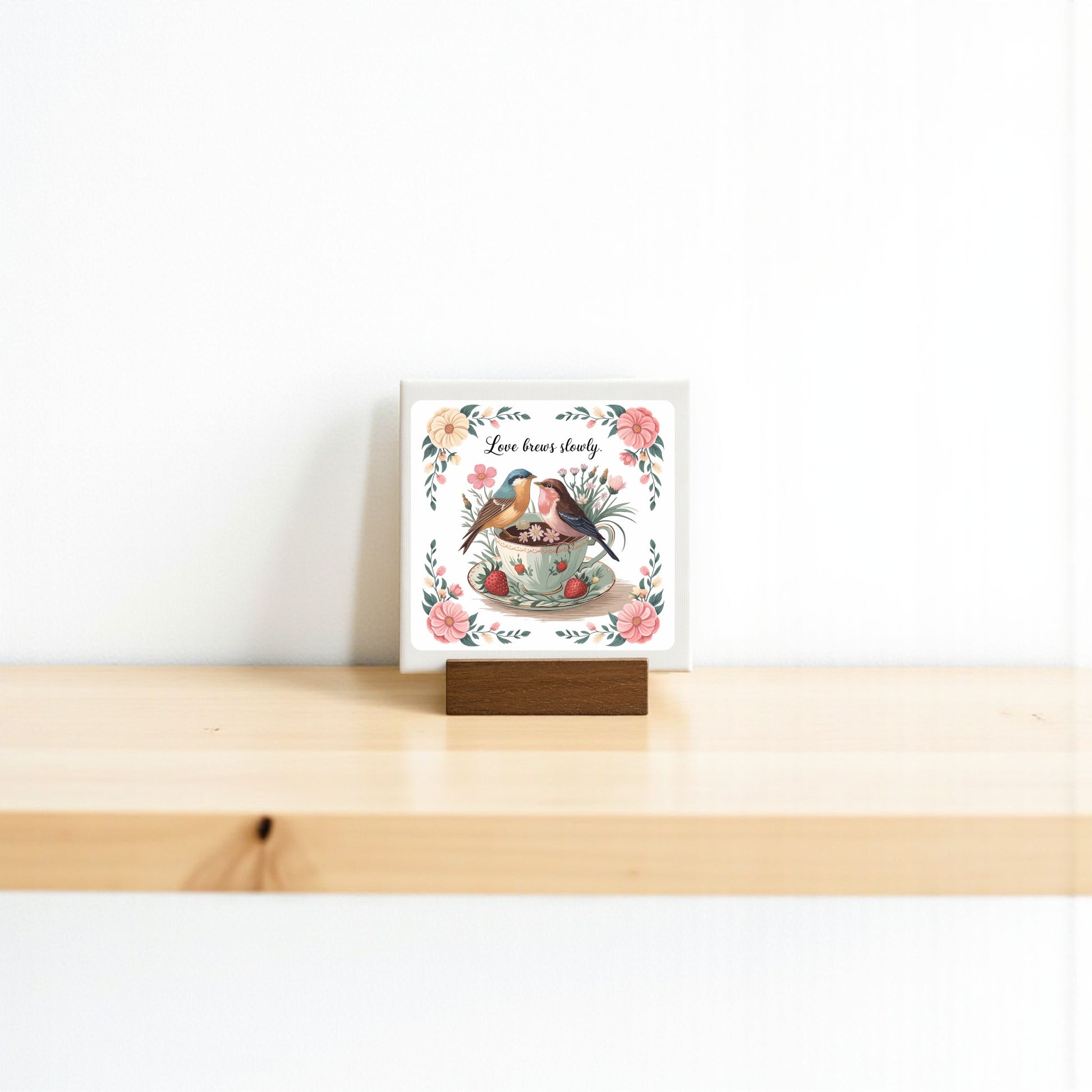 The Love Brews Slowly Mini Canvas by Mirabilia Boutique features two birds in a floral teacup with "Love lives here." Displayed on a wooden stand, this 4x4 romantic art is perfect cottagecore decor for a cozy touch.