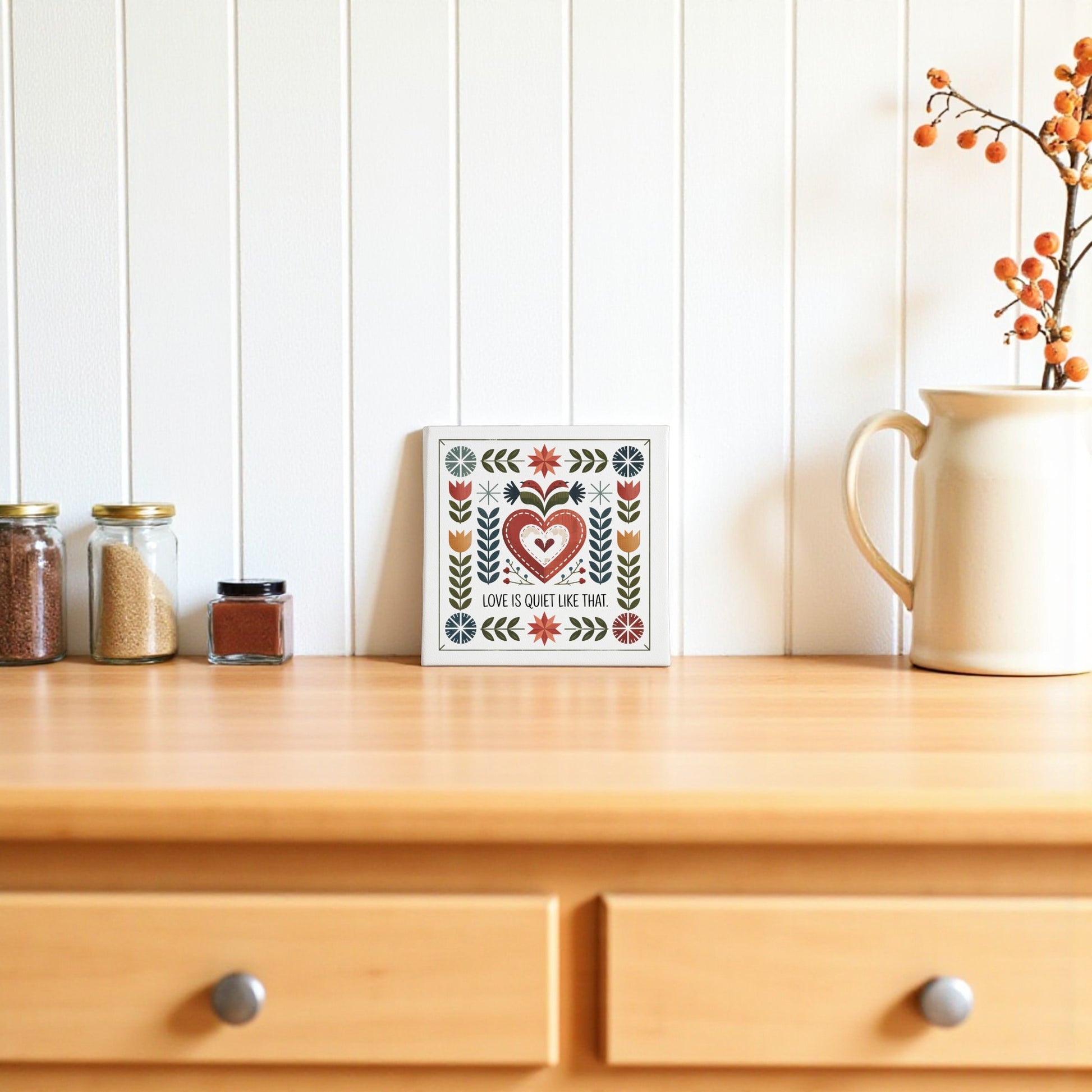 The Love Is Quiet Like That Canvas by Mirabilia Boutique, featuring folk art and a heartwarming quote, adds a charming and cozy touch to home decor on a white paneled wall.