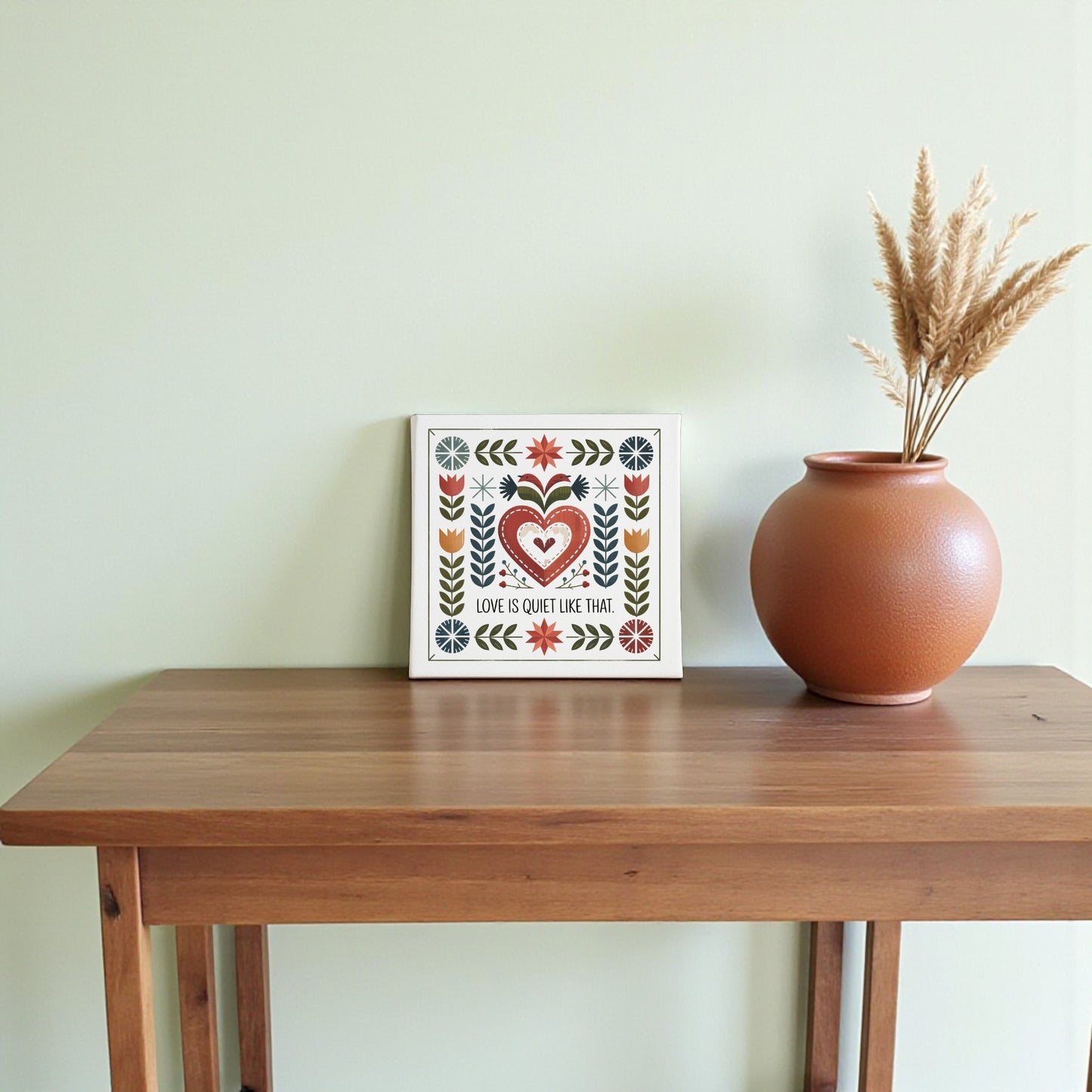 The Mirabilia Boutique "Love Is Quiet Like That" folk art canvas adds cozy charm to your home decor, displayed in a frame on a wooden table beside a brown vase with dried pampas grass against a light green wall.