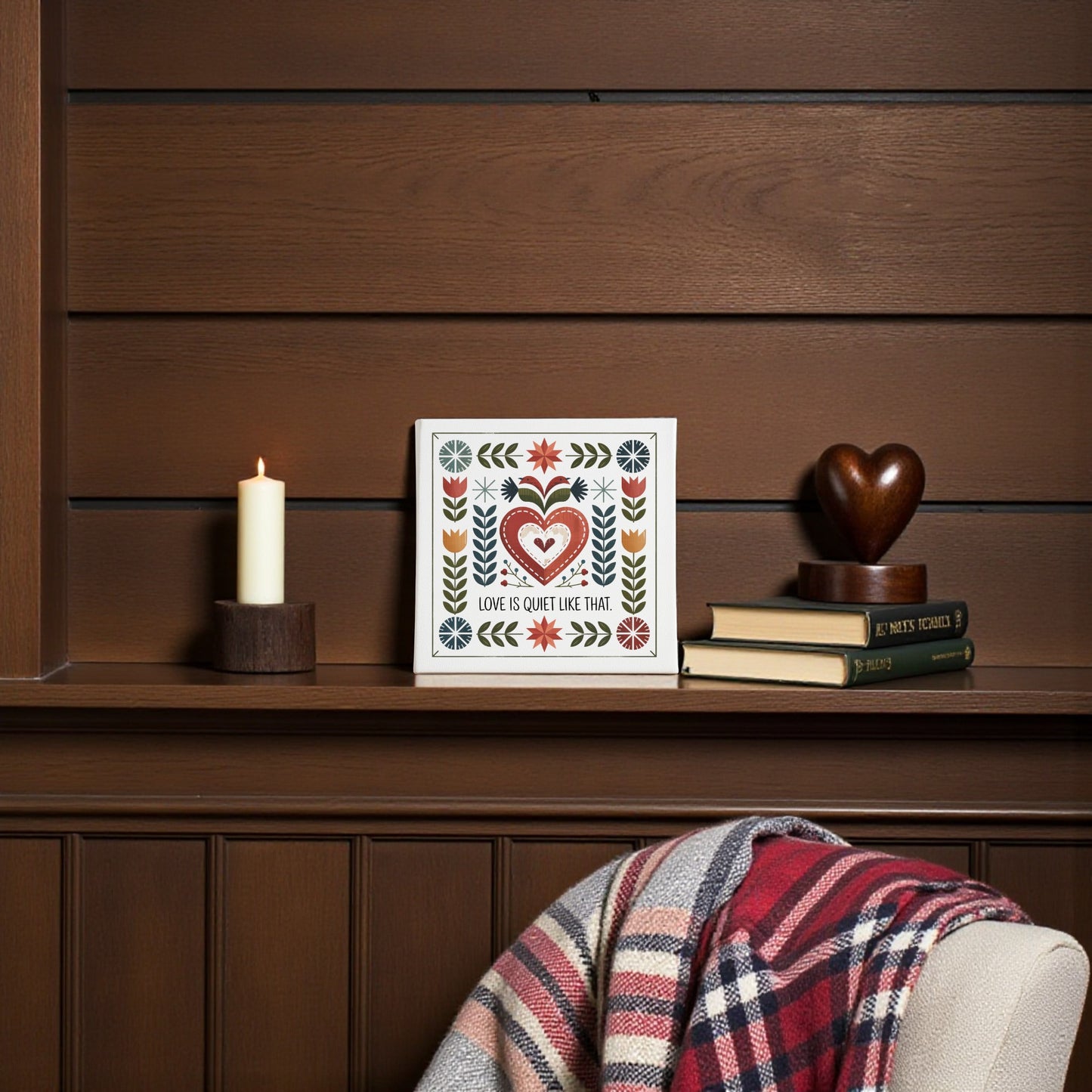 A cozy corner features Mirabilia Boutique's "Love Is Quiet Like That Canvas" with Nordic folk art hearts, surrounded by a lit candle, books, a wooden heart, and a plaid blanket, creating inviting folk art wall decor.