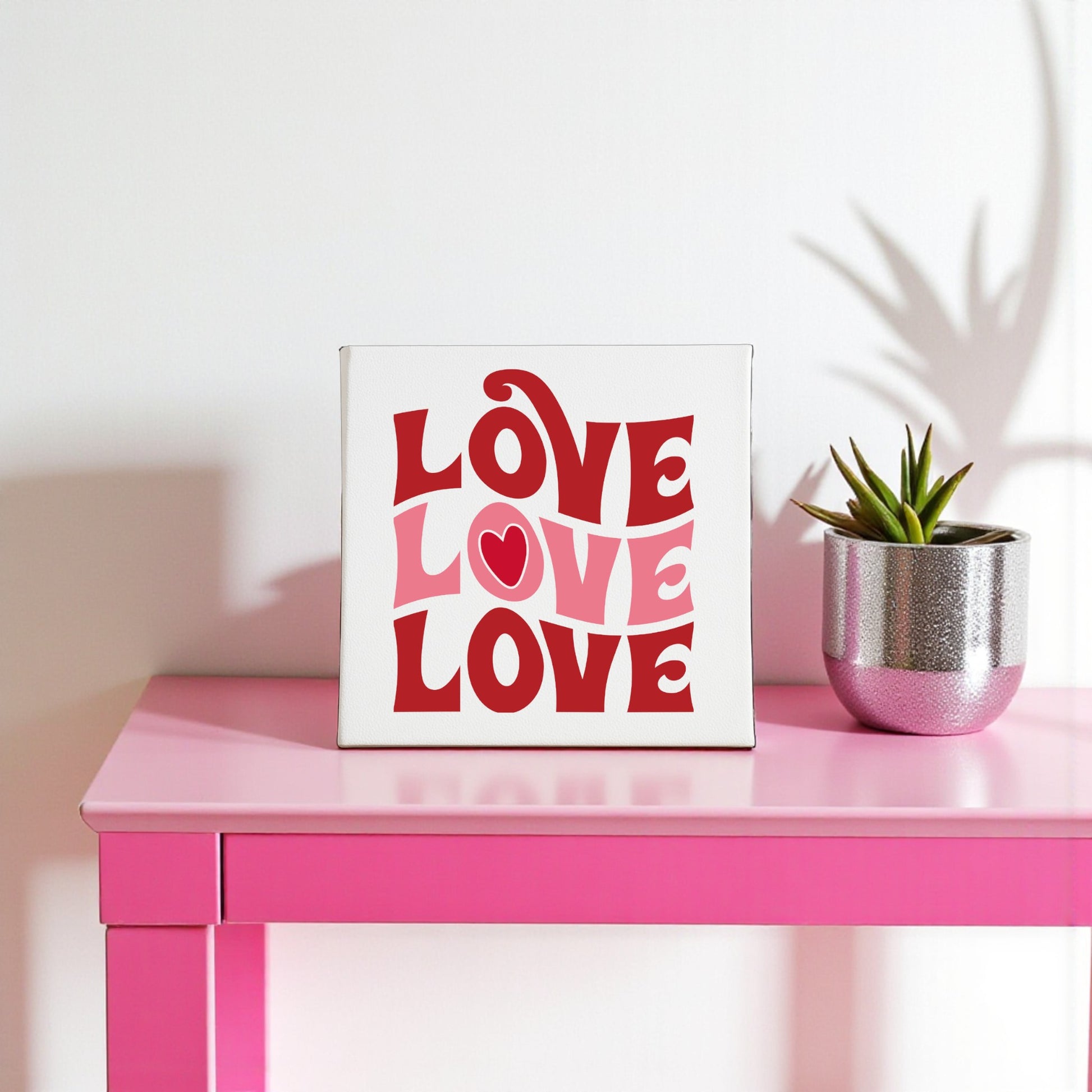 The Retro LOVE Canvas by Mirabilia Boutique features groovy red and pink lettering, displayed on a pink table with a potted plant—perfect Valentine’s Day decor for adding charm to your space.