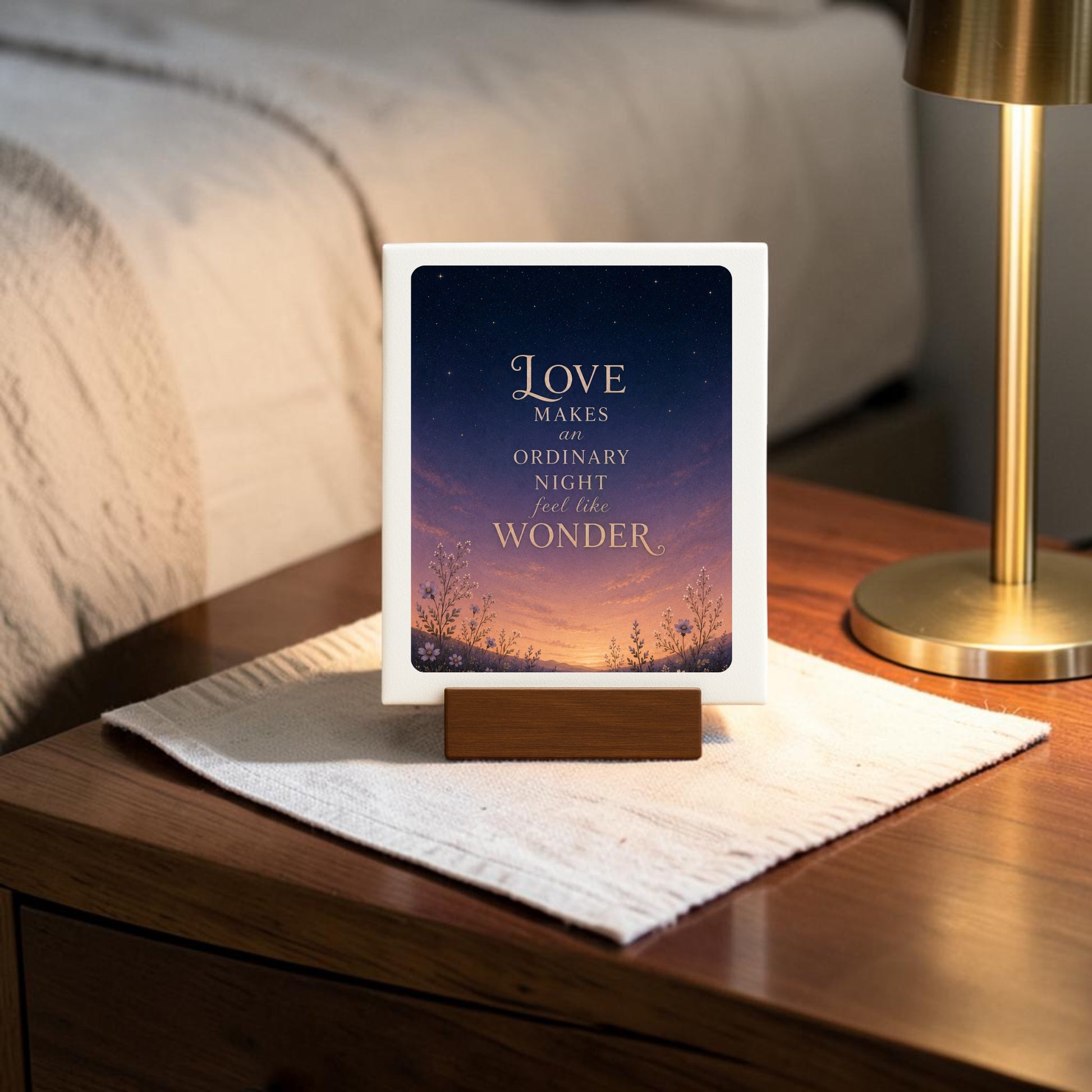 A Love Makes Wonder Romantic Mini Sign from Mirabilia Boutique’s Star & Solstice Collection sits atop a wooden nightstand, featuring the heartfelt message, “Love makes an ordinary night feel like wonder,” set against a starry sky and nature illustration. This celestial décor piece brings a romantic warmth beside a glowing lamp, with part of a bed visible in the background.