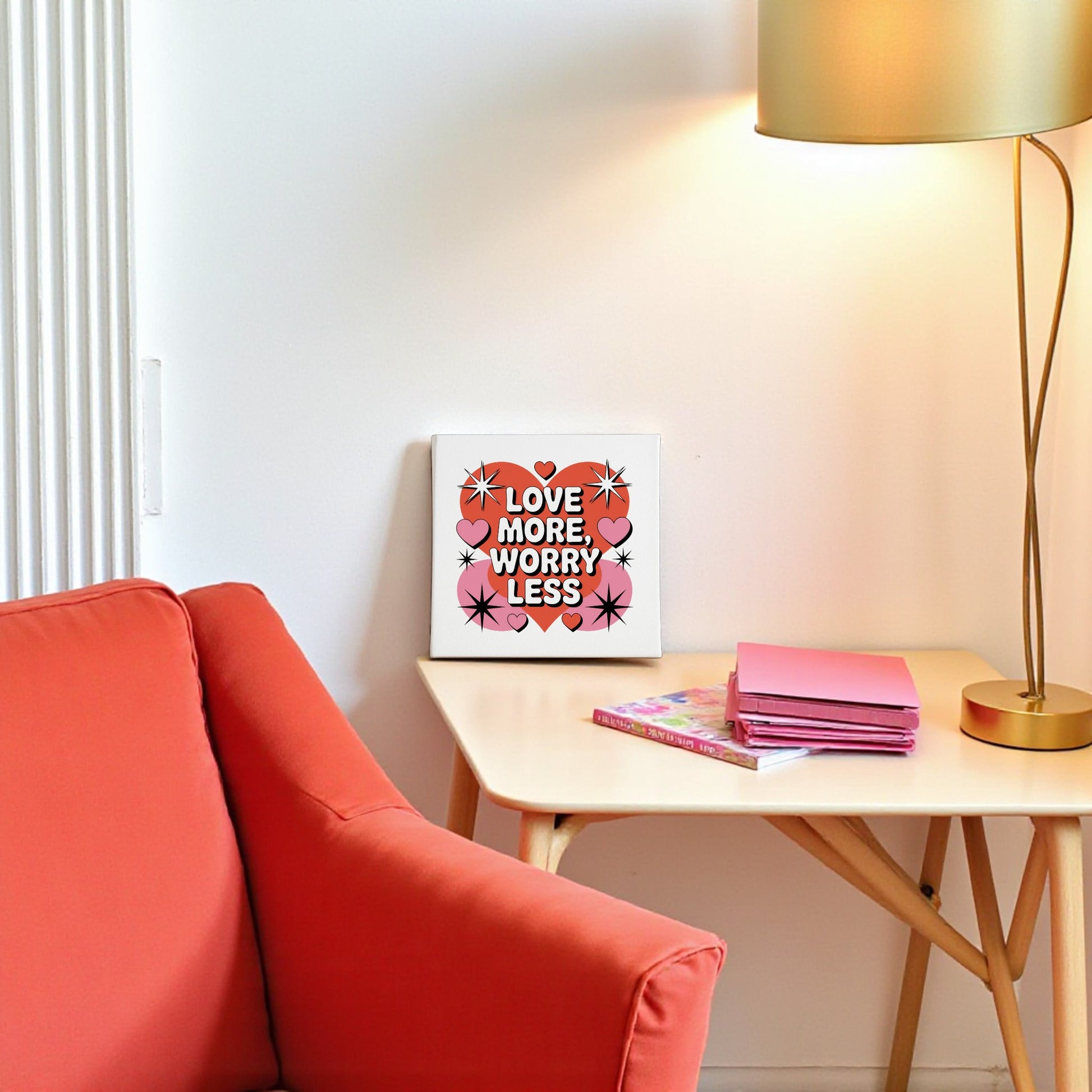 A cozy nook features a red armchair, a side table with pink notebooks, and a lamp. Displayed on the table is Mirabilia Boutique’s 8x8 "Love More Worry Less" Canvas, adding midcentury flair with its red hearts and stars.