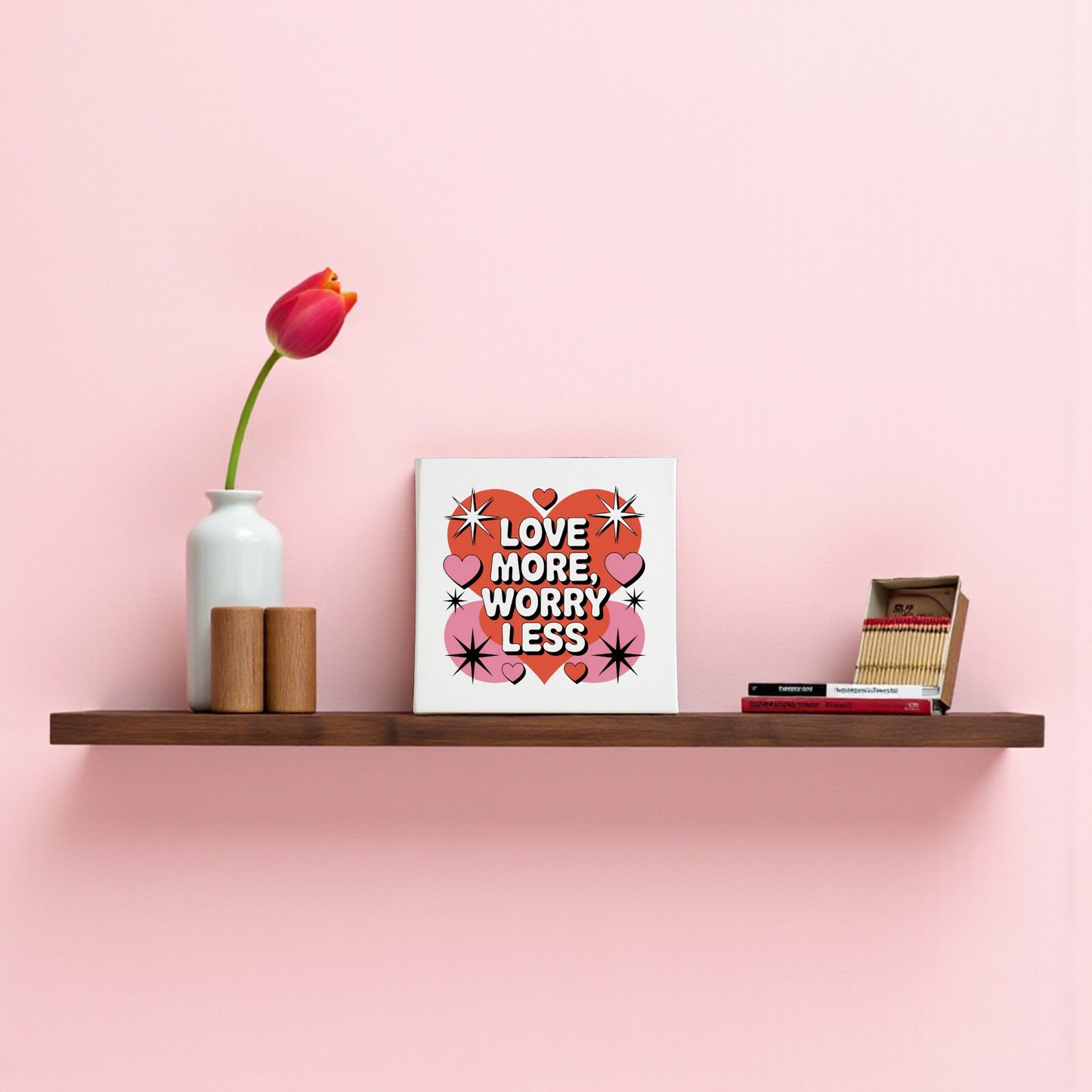 A pink wall features the Mirabilia Boutique "Love More Worry Less Canvas" (8x8, midcentury wall art), surrounded by a vase with a red tulip, two wooden containers, a heart motif, and a stack of books beside an open matchbox.