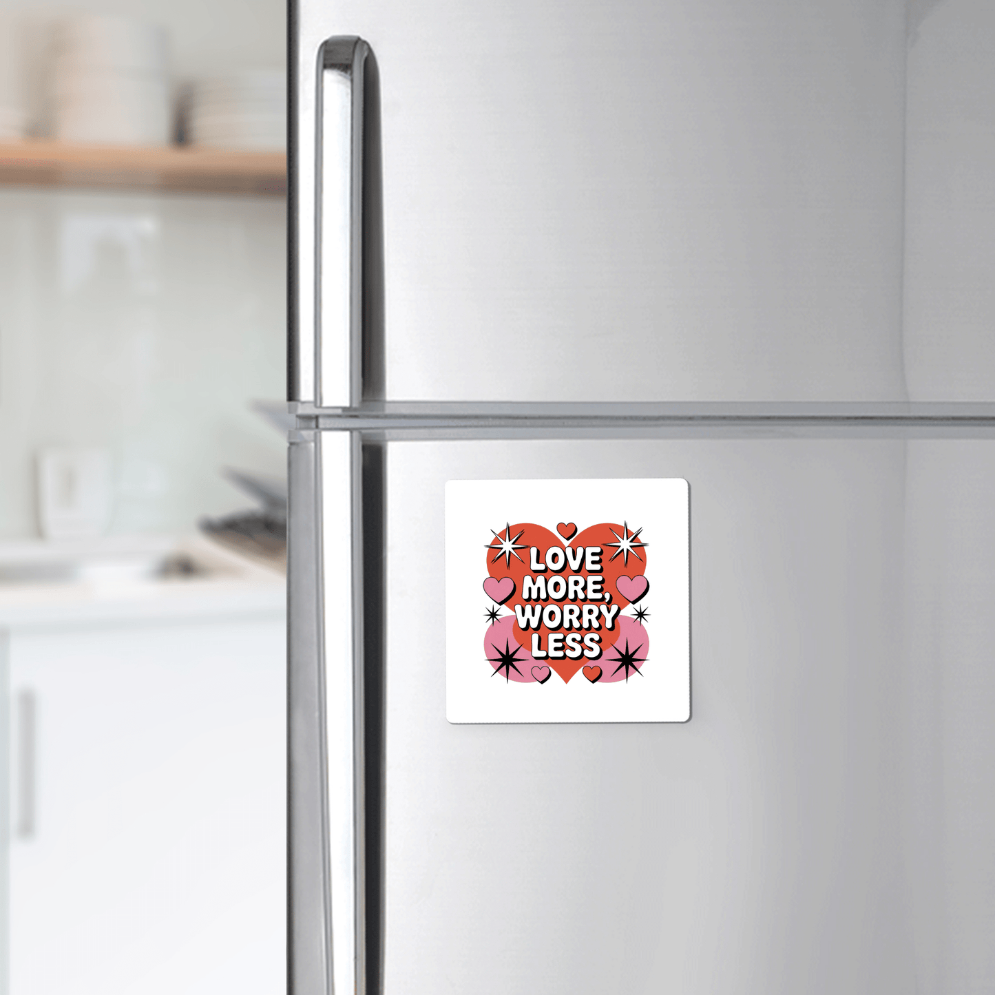 A silver fridge features the "Love More Worry Less Magnet | Retro Valentine Fridge Decor" by Mirabilia Boutique, decorated with bold letters, red hearts, and pink stars. Blurry white kitchen cabinets and dishes are visible behind.