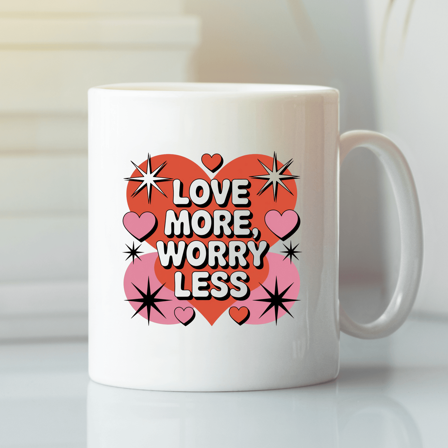 The Love More Worry Less Mug | Retro Valentine Coffee Cup by Mirabilia Boutique features red and pink hearts, black starbursts, and bold "LOVE MORE, WORRY LESS" text—perfect for Valentine’s Day or brightening your morning.