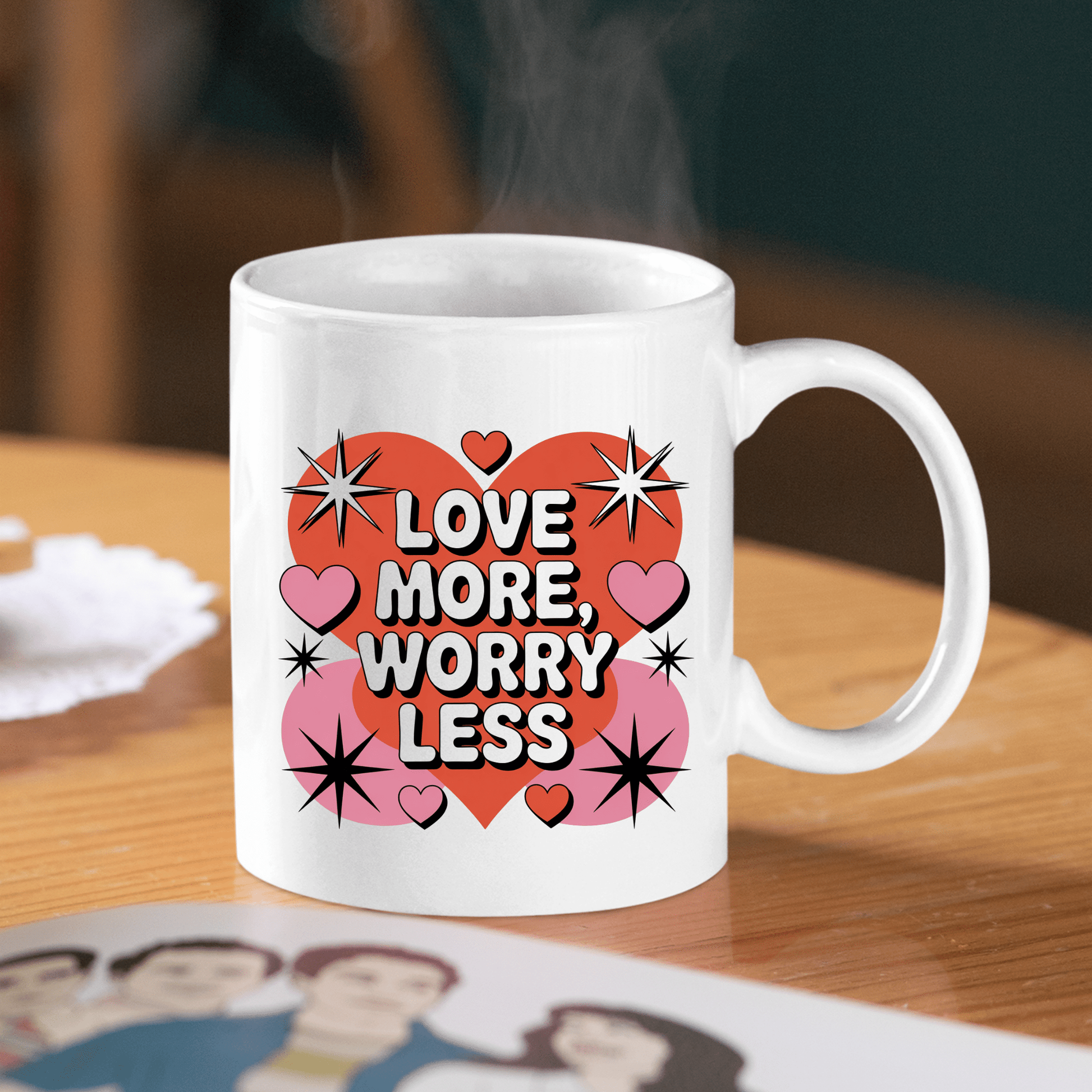 A Love More Worry Less Mug | Retro Valentine Coffee Cup by Mirabilia Boutique sits on a wooden table, steam rising as heart designs glow—making it a charming Valentine’s Day gift. A people-illustrated magazine rests in the foreground.