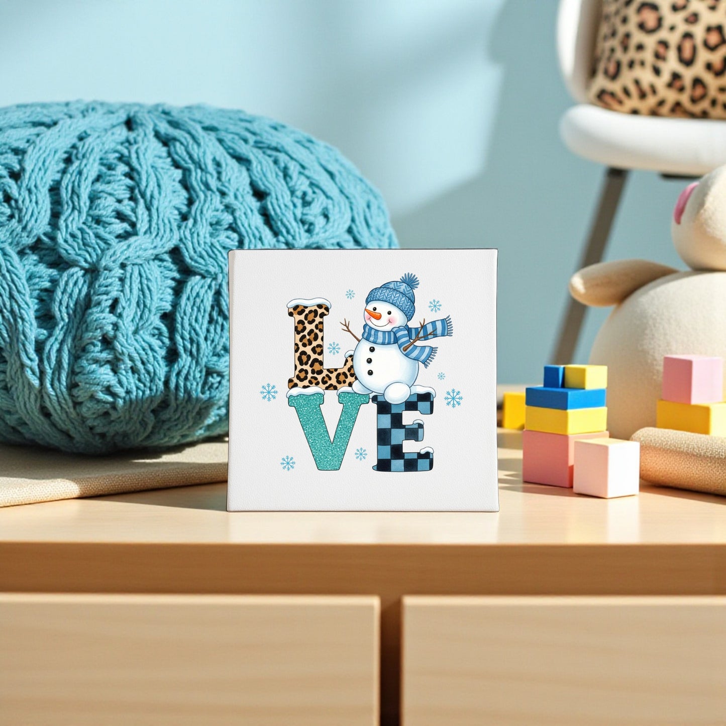 The LOVE Snowman Canvas Sign by Mirabilia Boutique spells “LOVE” with a snowman as the “O”—perfect winter or Valentine’s décor. Displayed with a blue knit blanket, toy blocks, plush toy, and a leopard-print chair.