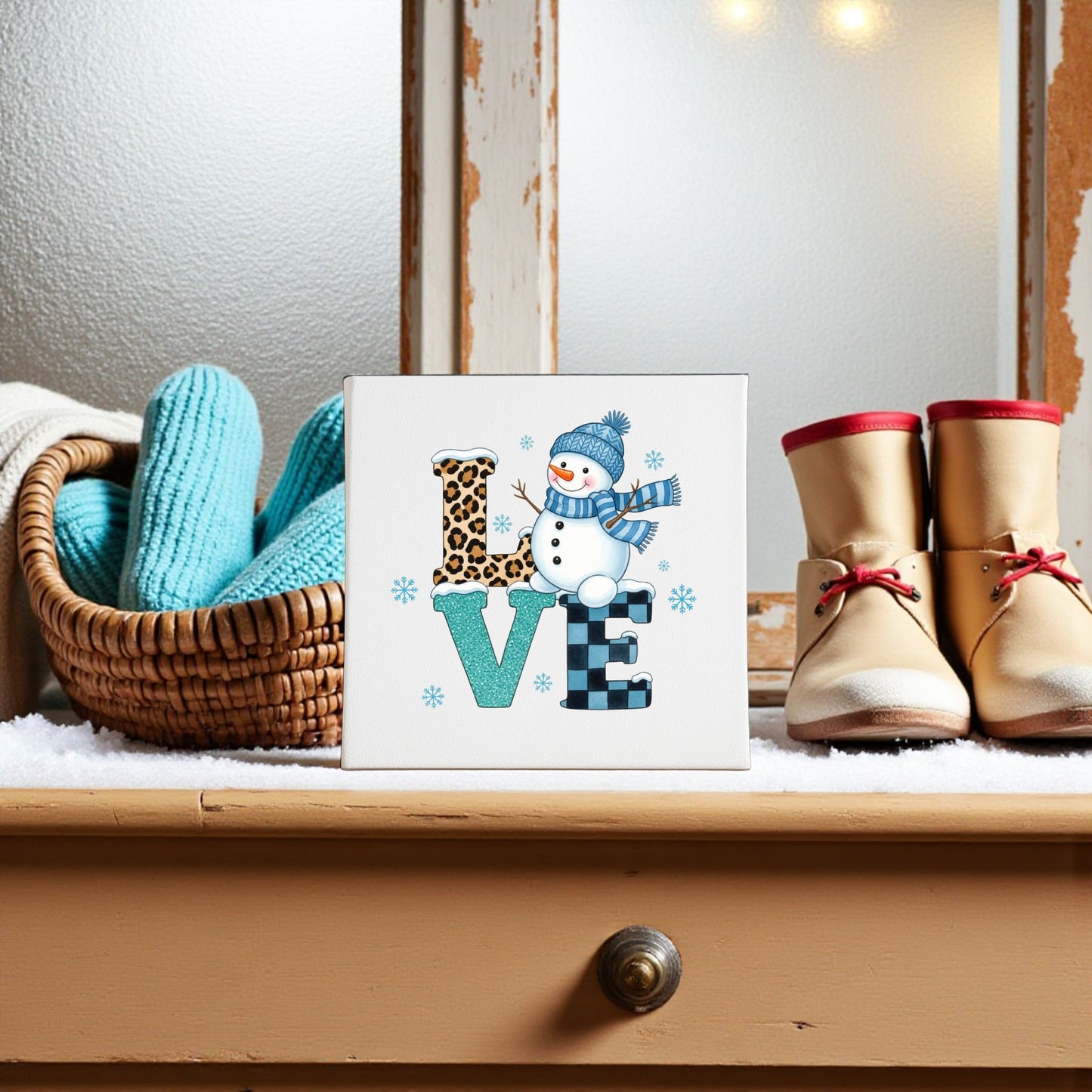 Create a cozy winter scene with Mirabilia Boutique's LOVE Snowman Canvas Sign, blue knit mittens in a basket, and boots on a snowy wooden table by a frosted window—perfect for winter or Valentine’s Day décor.
