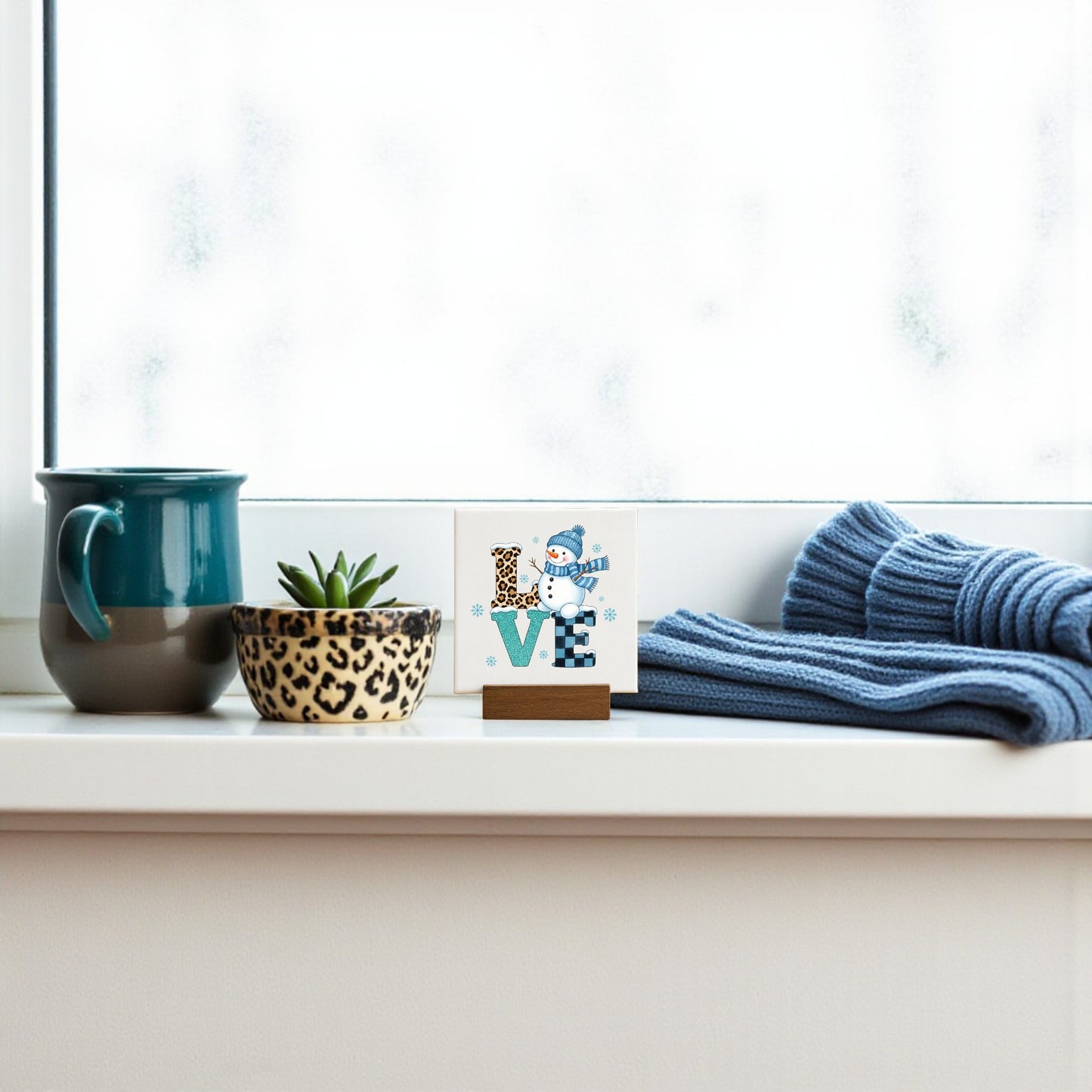 A windowsill with a teal mug, a leopard print pot holding a succulent, and Mirabilia Boutique’s Mini LOVE Snowman Sign creates cozy winter shelf decor against a frosted window backdrop.