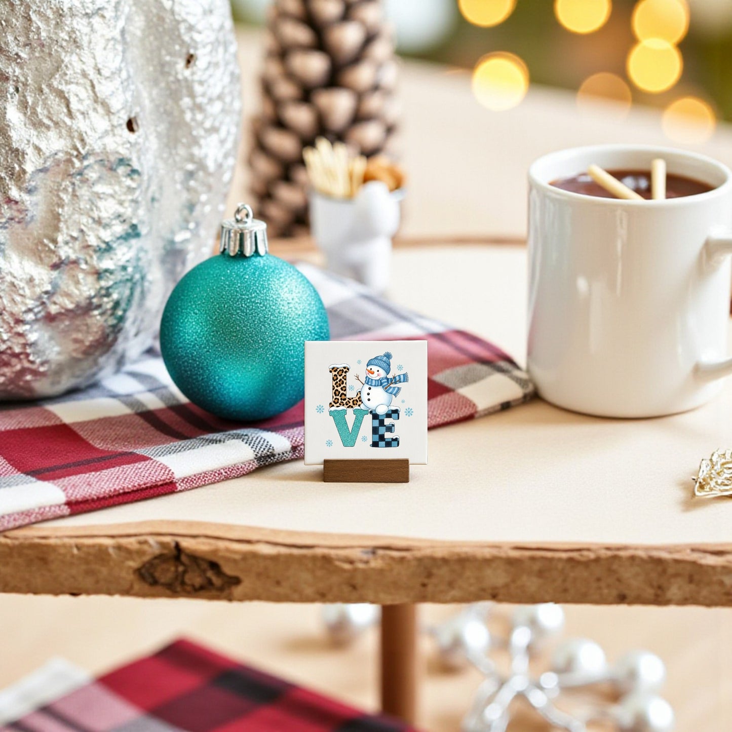 The Mini LOVE Snowman Sign by Mirabilia Boutique adds charming winter Valentine decor to your table, paired with a blue ornament, a cozy mug, and plaid napkin, softly lit by festive background lights.