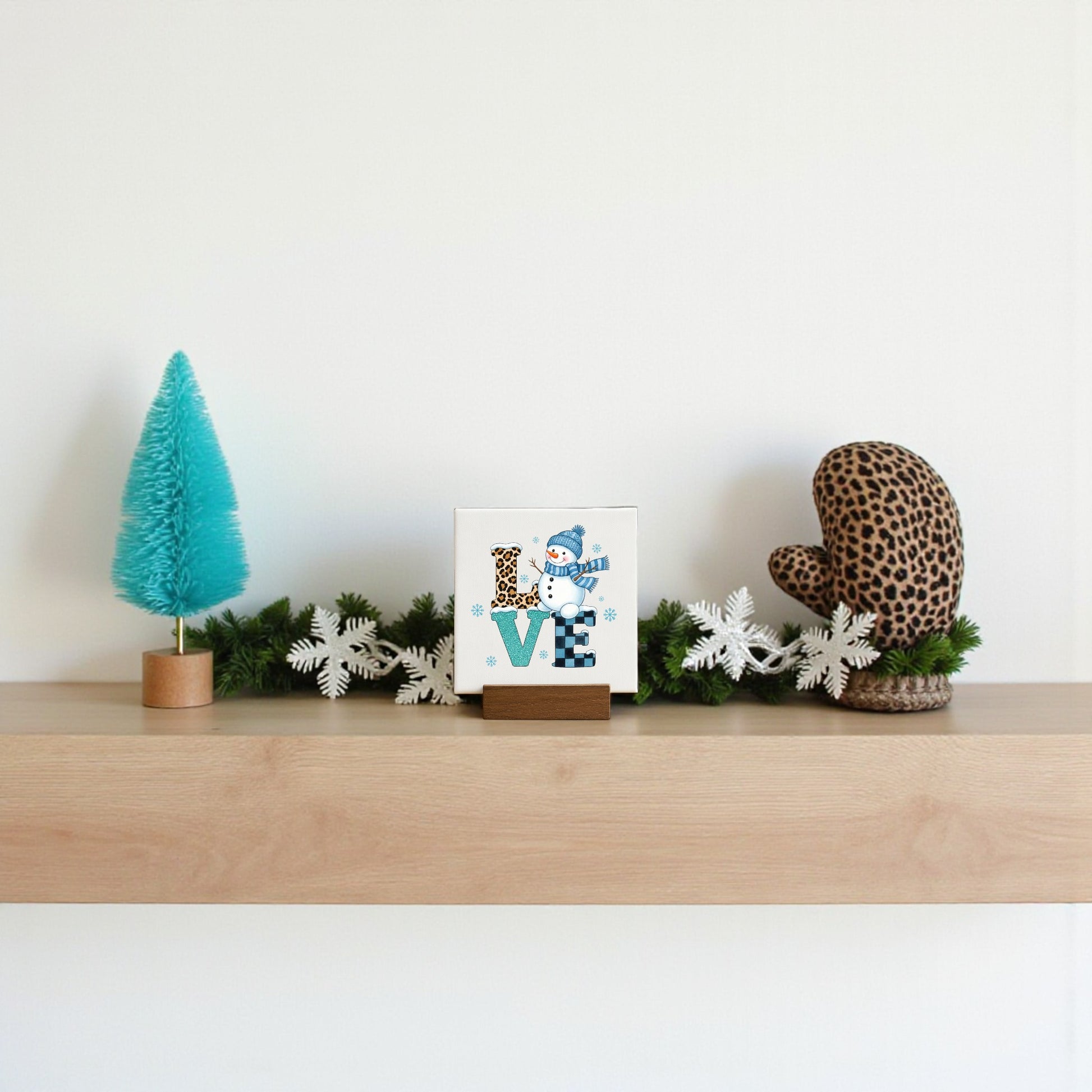A minimalist wooden shelf displays winter decor: a blue bottle-brush tree, a Mirabilia Boutique Mini LOVE Snowman Sign, white faux pine garland, and a brown leopard-print mitten against a plain white wall.