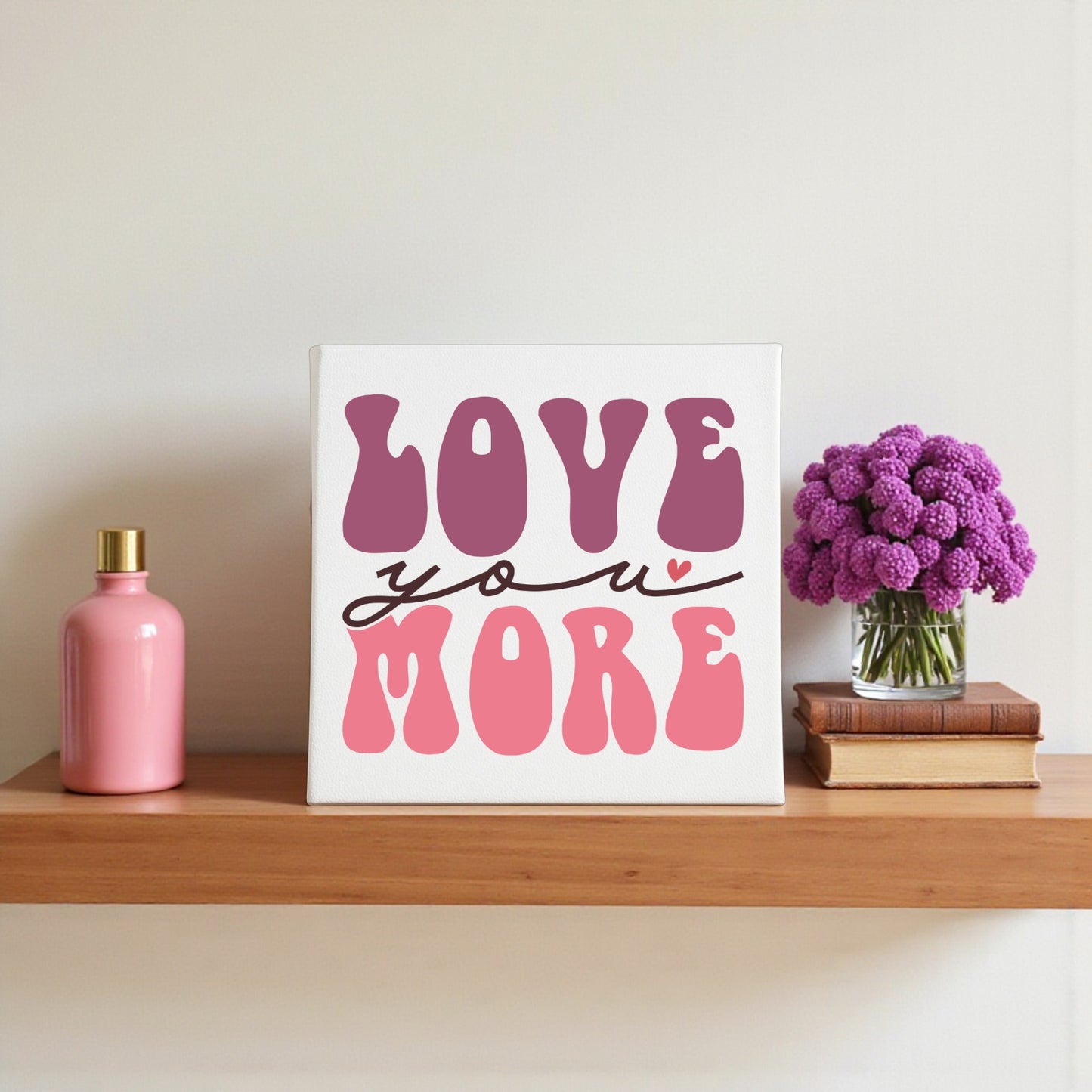 A wooden shelf displays a pink vase, Mirabilia Boutique’s Love You More 8x8 Canvas | Retro Valentine Wall Art, two stacked books, and a bouquet of purple flowers in a glass jar.