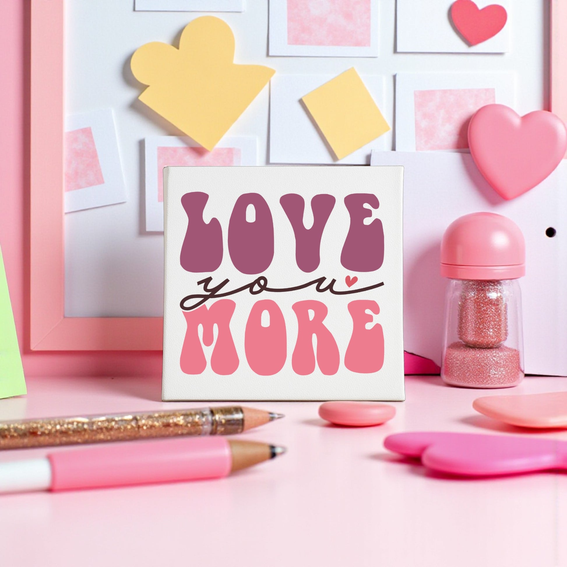 A pink, heart-themed desk features the "Love You More 8x8 Canvas | Retro Valentine Wall Art" by Mirabilia Boutique in bold retro pink and purple, with groovy wall art, sticky notes, pens, and a framed board as accents.
