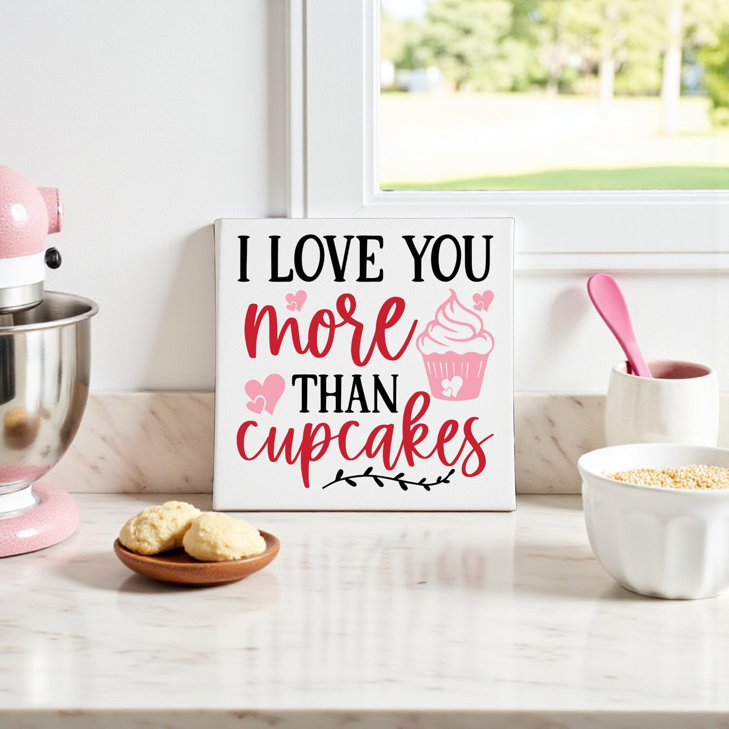 A kitchen counter displays Valentine’s decor, including the Mirabilia Boutique “I Love You More Than Cupcakes” 8x8 canvas sign, a stand mixer, cookies on a plate, and a mixing bowl with a pink spoon by the window.
