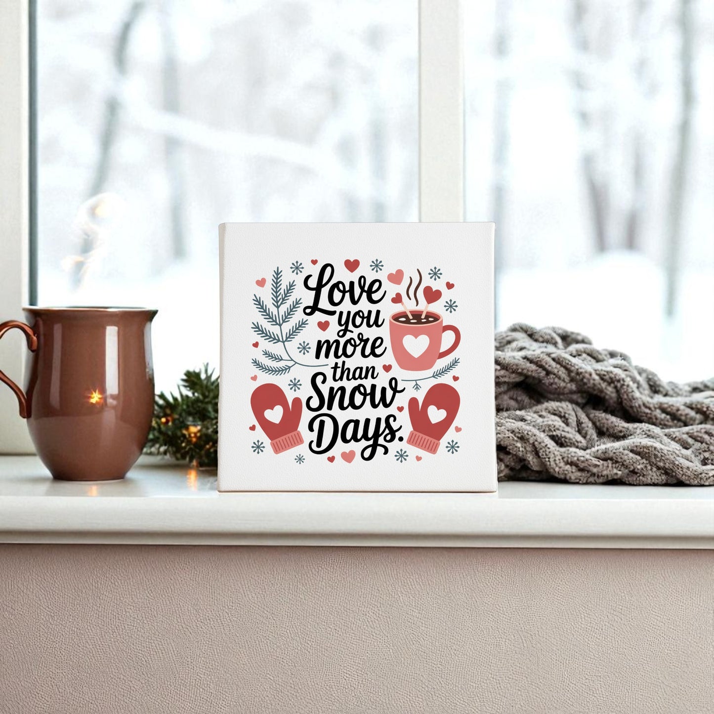 A ceramic mug and a knitted gray blanket sit on a windowsill beside Mirabilia Boutique’s "Love You More Than Snow Days" print—a charming winter wall art gift—while snowy trees outside set the perfect festive scene.