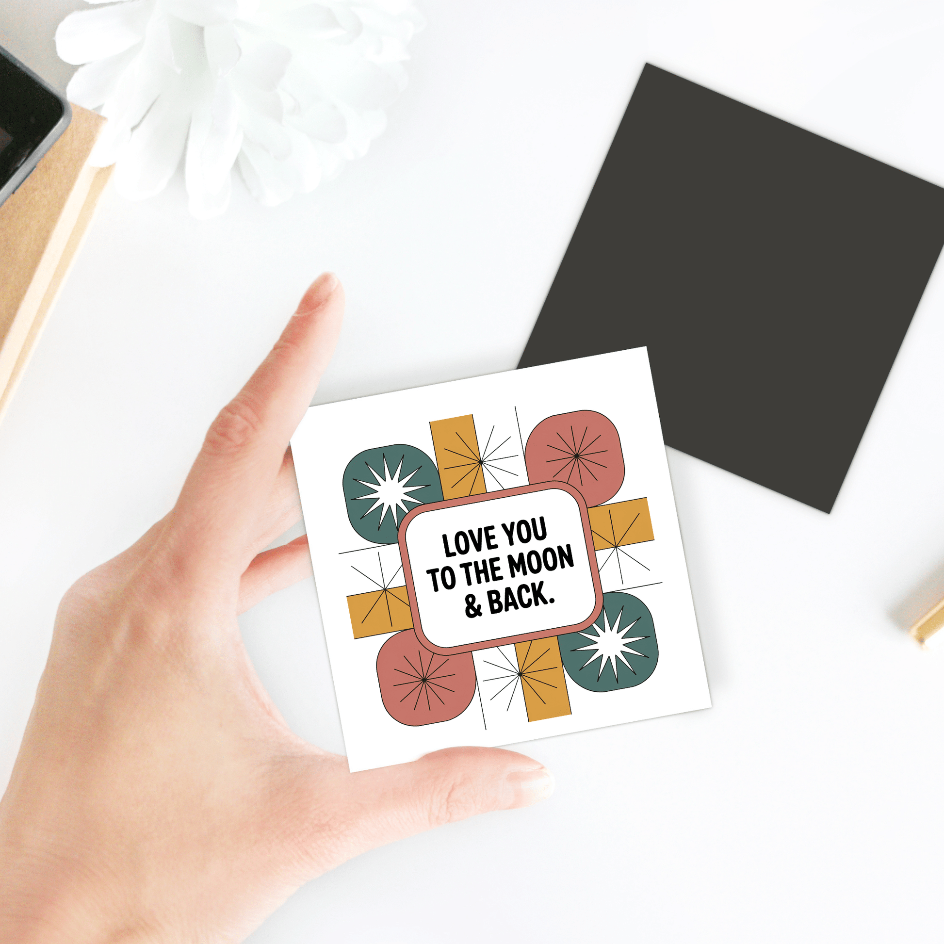A hand holds the Retro Love Magnet by Mirabilia Boutique, featuring "LOVE YOU TO THE MOON & BACK" in colorful retro style. In the background, a black square and a white flower rest on the white table.