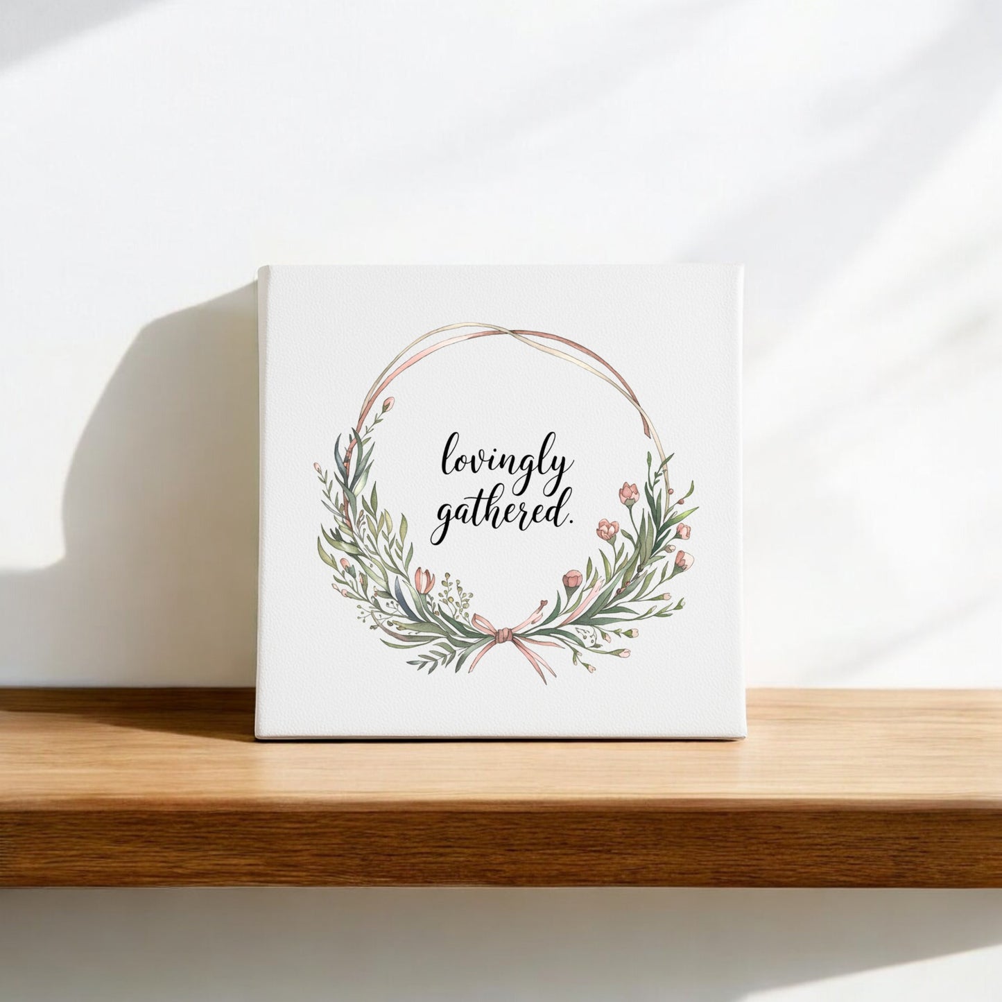 The Mirabilia Boutique "Lovingly Gathered Canvas – 8x8 Botanical Wreath Wall Art" features green leaves and pink flowers on a white square canvas, encircling “lovingly gathered” for a charming touch to any home decor.