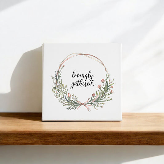 The Mirabilia Boutique "Lovingly Gathered Canvas – 8x8 Botanical Wreath Wall Art" features green leaves and pink flowers on a white square canvas, encircling “lovingly gathered” for a charming touch to any home decor.