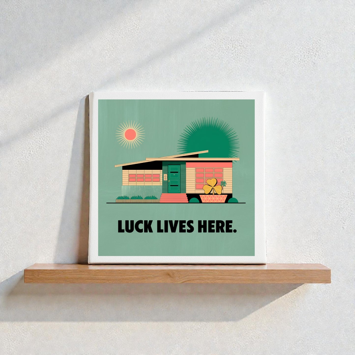 The Luck Lives Here Canvas Sign – Midcentury Home Art by Mirabilia Boutique sits on a wooden shelf, showcasing a modern house, two sunbursts, and bold lettering against a textured white wall.