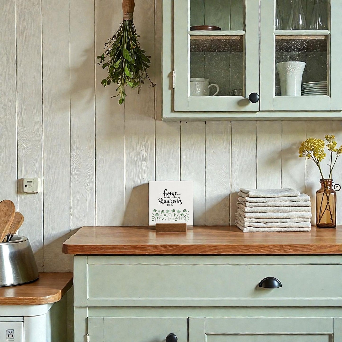 Kitchen scene with pale green cabinets, wooden countertop, stacked towels, herbs hanging on the wall, a vase of yellow flowers, and Mirabilia Boutique’s "Home Is Where the Shamrocks Grow" mini sign—perfect Irish home decor charm.