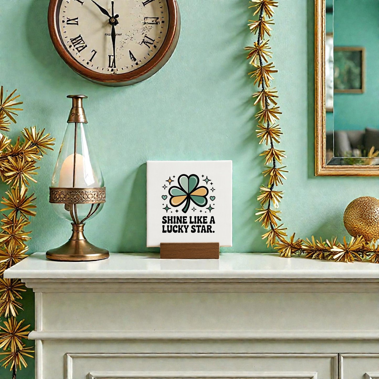 A mantel adorned with gold star garland, a lamp, and the Mirabilia Boutique “Shine Like a Lucky Star” 4x4 Canvas Sign sits beneath a clock, beside a gold ball and framed mirror on a green wall for St. Patrick’s Day flair.