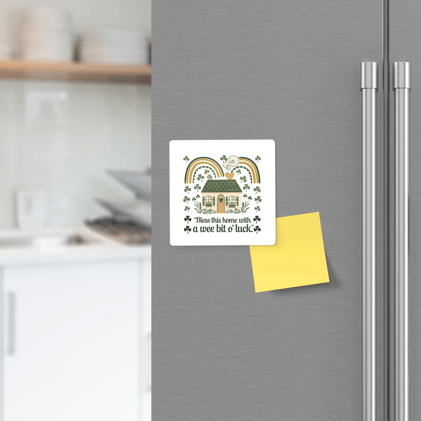 A Mirabilia Boutique Wee Bit o’ Luck Irish Home Magnet is on the fridge beside a blank yellow sticky note, with kitchen shelves in the background. The magnet features a cottage, rainbow, clovers, and the phrase "Bless this home with a wee bit o' luck.