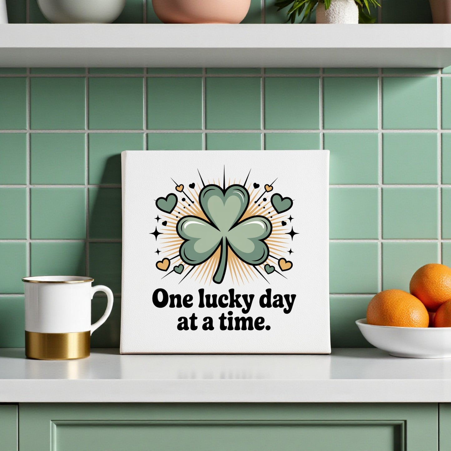 A kitchen with green tiled walls displays the Mirabilia Boutique's “One Lucky Day at a Time Canvas,” decorated with a shamrock, hearts, and sparkles. A mug and a bowl of oranges sit nearby on the counter.