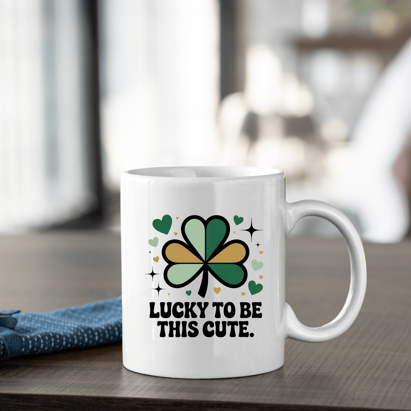 The Mirabilia Boutique "Lucky to Be This Cute Shamrock Mug" sits on a wooden table, decorated with green and gold hearts and clovers, bold black text, and a blue napkin beside it.