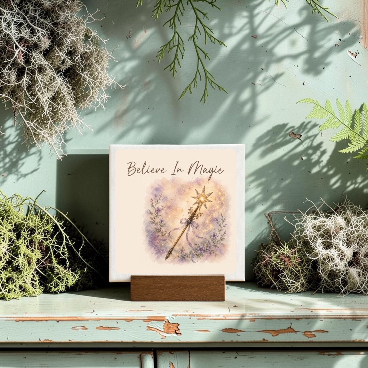 The Believe In Magic Mini Canvas Sign by Mirabilia Boutique features a fairy wand design and the inspiring phrase "Believe In Magic." Displayed on a wooden stand surrounded by moss and ferns atop a rustic, pale green surface, this piece is bathed in sunlight that casts soft shadows—creating an enchanting atmosphere perfect for watercolor floral decor.