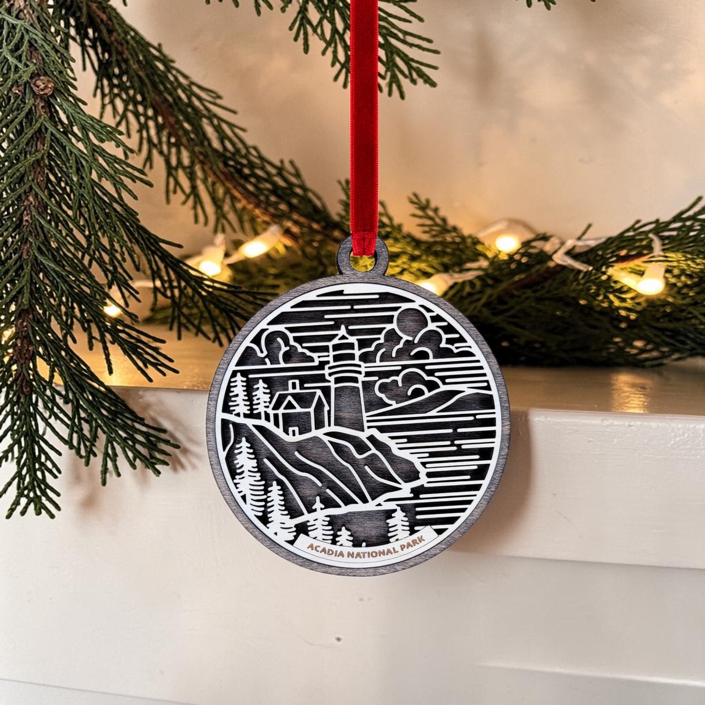 The Acadia National Park Line Art Ornament by Mirabilia Boutique features a charming lighthouse scene and "Acadia National Park" inscribed on it. Hanging from a red ribbon on a pine branch amid string lights and festive greenery, this round ornament makes the perfect Maine coast decor for your holiday celebrations.