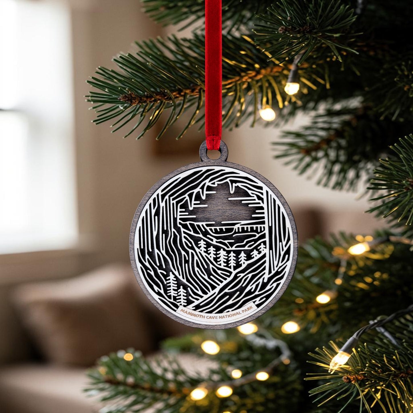 The Mammoth Cave National Park Line Art Christmas Ornament by Mirabilia Boutique is a handcrafted black and white wooden keepsake featuring a cave and forest design. It hangs from a red ribbon on a lit Christmas tree branch, set against a softly blurred indoor background.