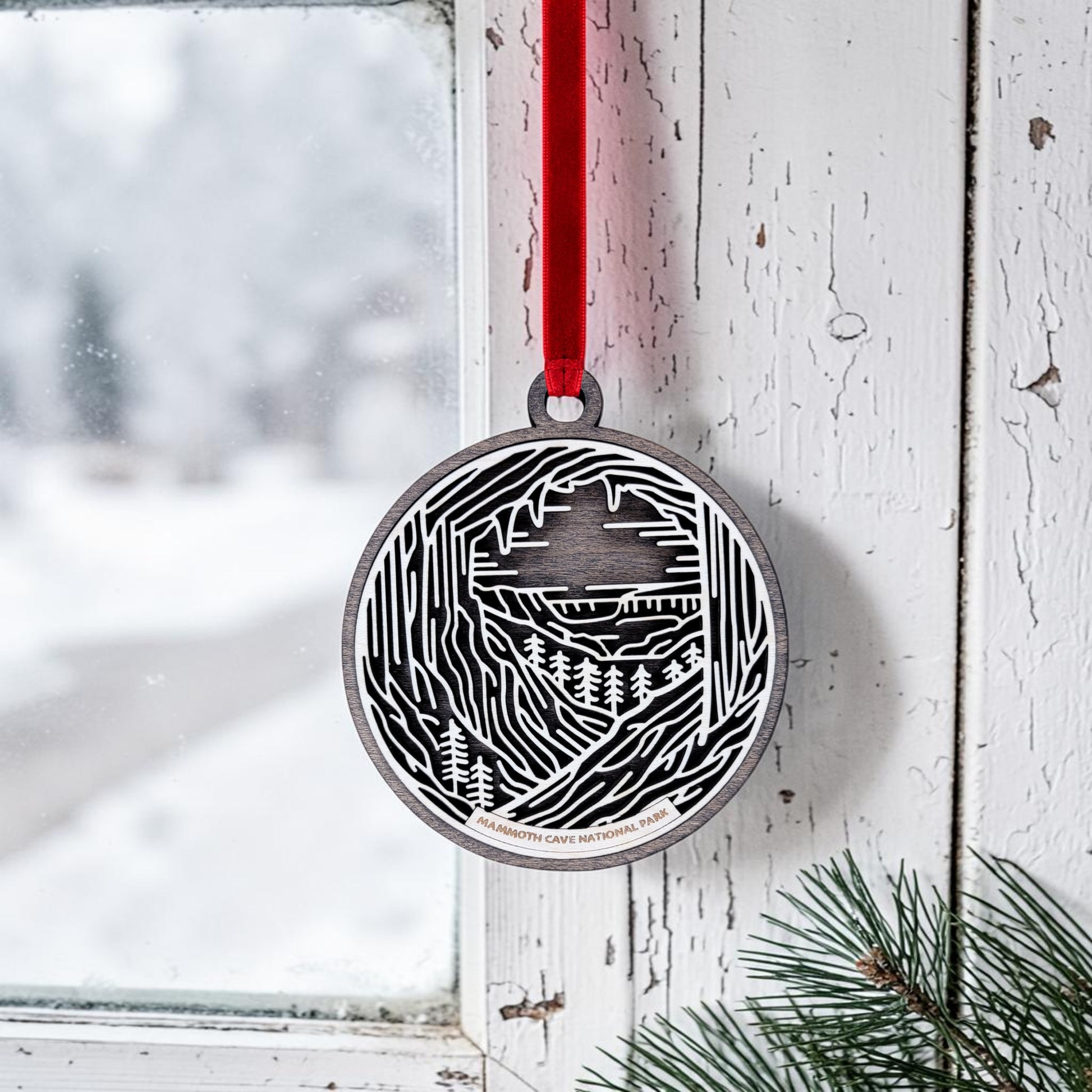 Celebrate the serene beauty of Mammoth Cave National Park with the Mammoth Cave National Park Line Art Christmas Ornament by Mirabilia Boutique. This handcrafted metal ornament, adorned with an elegant mountain and cave design, hangs from a red ribbon on a window frame. Snowy scenery is visible outside, while pine branches rest on the windowsill, capturing the peaceful spirit of the park during wintertime.