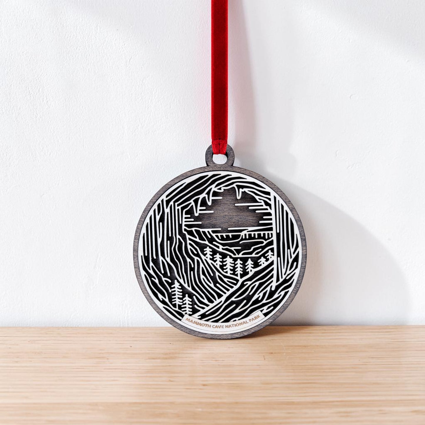 The Mammoth Cave National Park Line Art Christmas Ornament by Mirabilia Boutique is a round wooden keepsake featuring an abstract black and white engraving of a cave and trees. Accented with a red ribbon, this handcrafted ornament rests on a wooden surface against a white wall, beautifully capturing the essence of Mammoth Cave National Park.