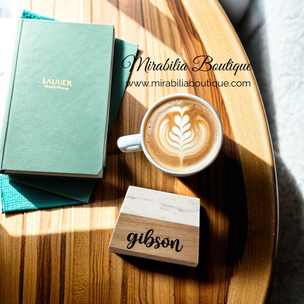 A wooden table holds a latte with a heart design, a green notebook labeled "Lauer," and a rectangular wooden box containing an engraved coaster from the Custom Engraved Marble and Acacia Coaster Set by Mirabilia Boutique. Sunlight casts shadows across the items.