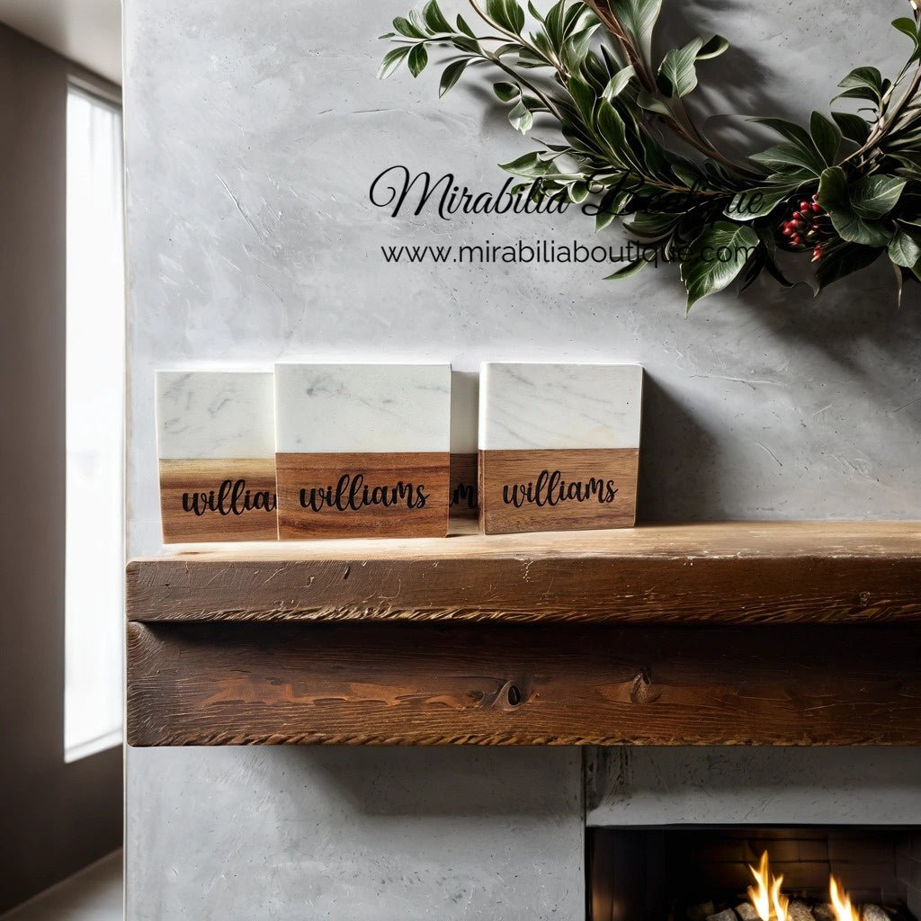 In a minimalist room, a wooden mantle showcases three Engraved Marble and Acacia Coasters from Mirabilia Boutique, each personalized with the name "Williams." Above, greenery and a website URL adorn the light gray textured wall. The cozy ambiance is enhanced by a partial view of the fireplace on the right.