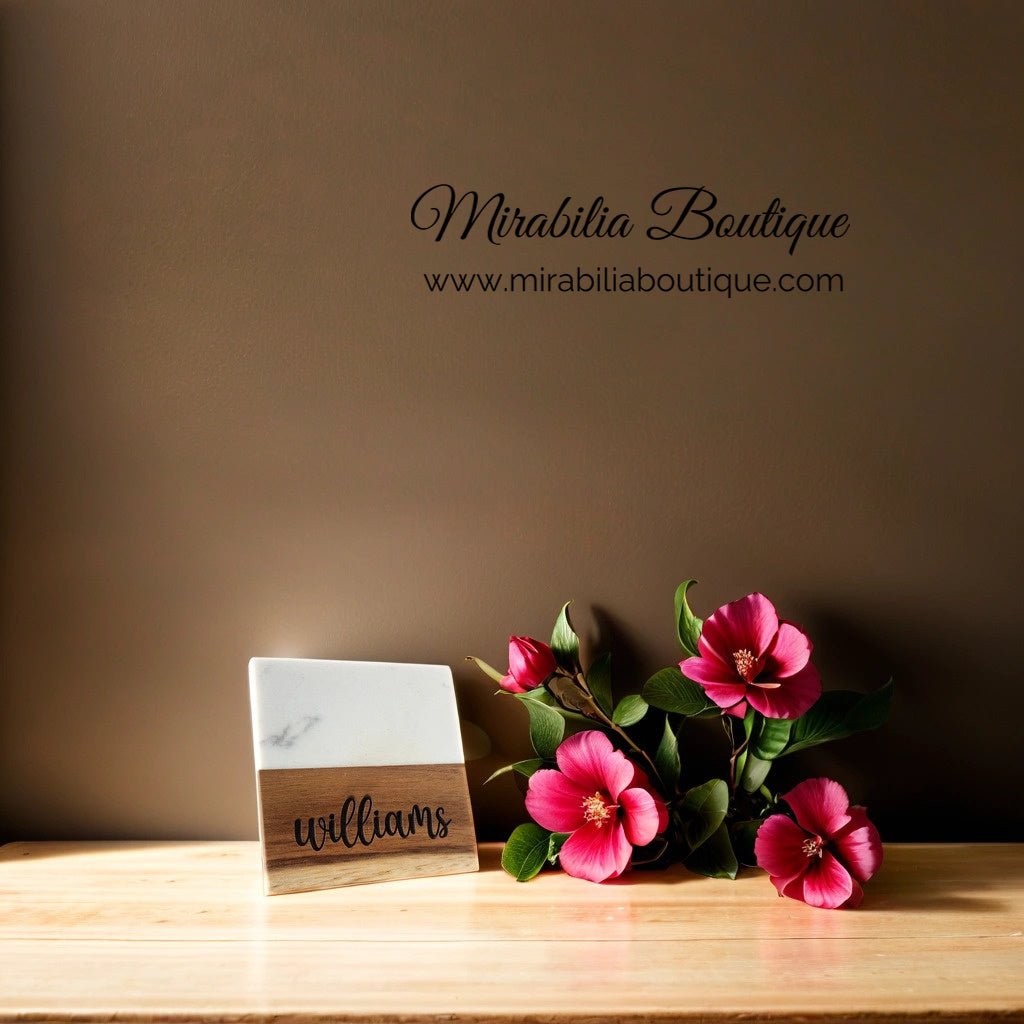 A sign displaying the word "williams" is placed on a wooden surface next to bright pink flowers, alongside a Custom Engraved Marble and Acacia Coaster Set. The wall in the background showcases the brand "Mirabilia Boutique" along with their website address, "www.mirabiliaboutique.com.