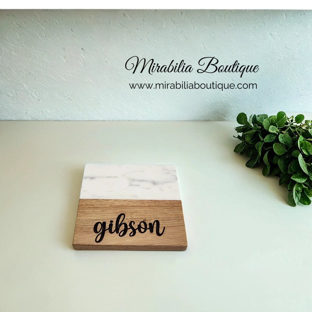 A Custom Engraved Marble and Acacia Coaster Set, featuring the name "gibson" on a section crafted from Acacia wood and enhanced with a white marble design, sits elegantly beside green foliage. In the background, the brand "Mirabilia Boutique" and its website link add a touch of sophistication to the scene.