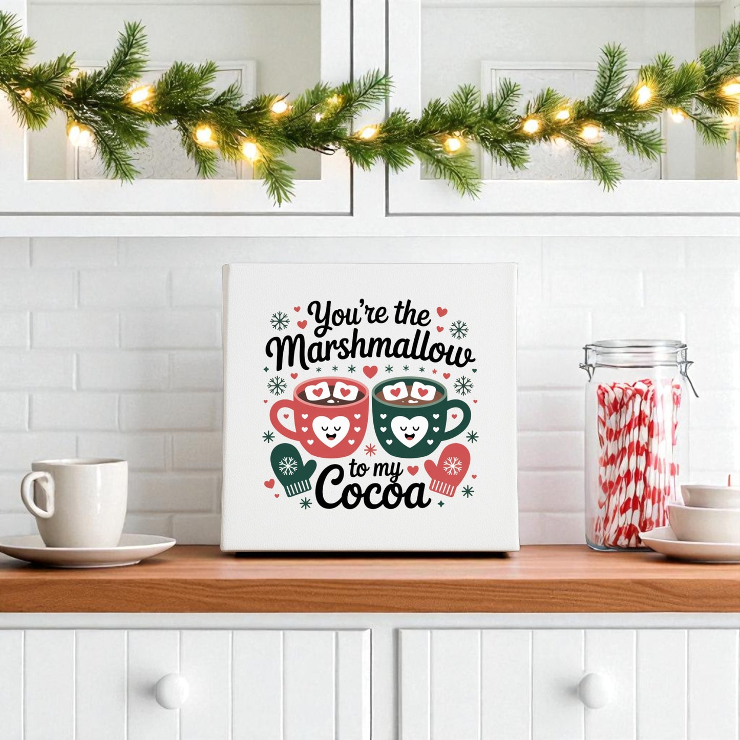 Create a cozy kitchen scene with the Mirabilia Boutique Marshmallow to My Cocoa Sign (8x8 Winter Canvas Art), featuring two smiling cocoa mugs, plus lighted garland, mugs, candy canes, and festive white shelf decor.