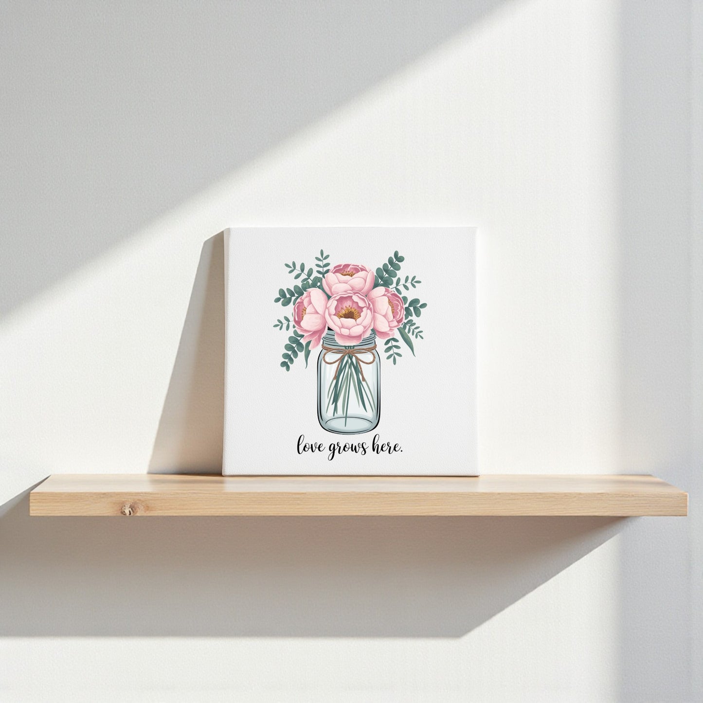 The Love Grows Here Floral Canvas Wall Art by Mirabilia Boutique rests on a wooden shelf, with blush peonies design illuminated by sunlight, casting gentle shadows on the white wall behind.