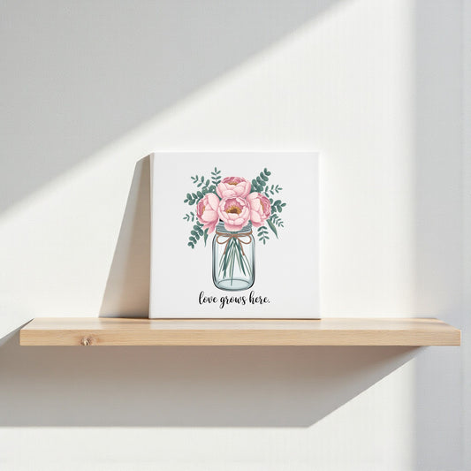 The Love Grows Here Floral Canvas Wall Art by Mirabilia Boutique rests on a wooden shelf, with blush peonies design illuminated by sunlight, casting gentle shadows on the white wall behind.