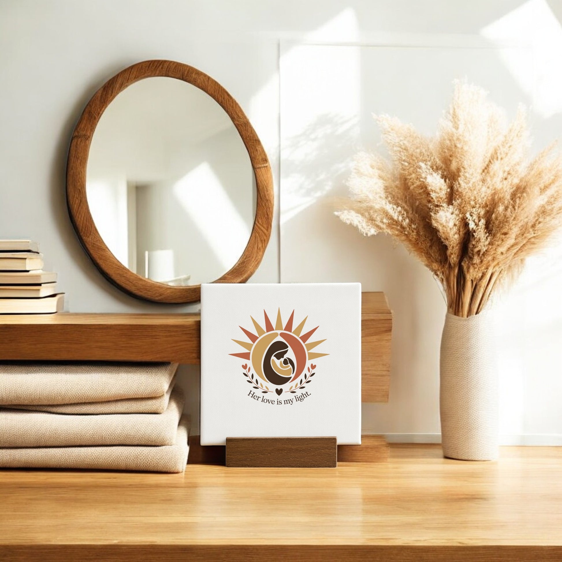 A wooden dresser displays stacked books, a round mirror, a white vase with dried pampas grass, and Mirabilia Boutique’s "Her Love Is My Light" Mini Sign – Mother’s Day Canvas. Sunlight brightens this heartfelt mother’s gift scene.