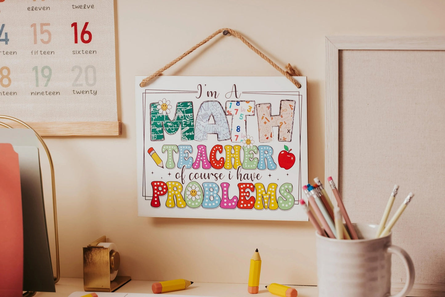 A Mirabilia Boutique sign hung on the wall reads, "I'm a Math Teacher: Humorous Problems Hanging Sign," surrounded by vibrant math-themed decor. Nearby, there's a mug filled with pencils, colorful chalk, and a section of a wall calendar displaying month numbers in soft hues.
