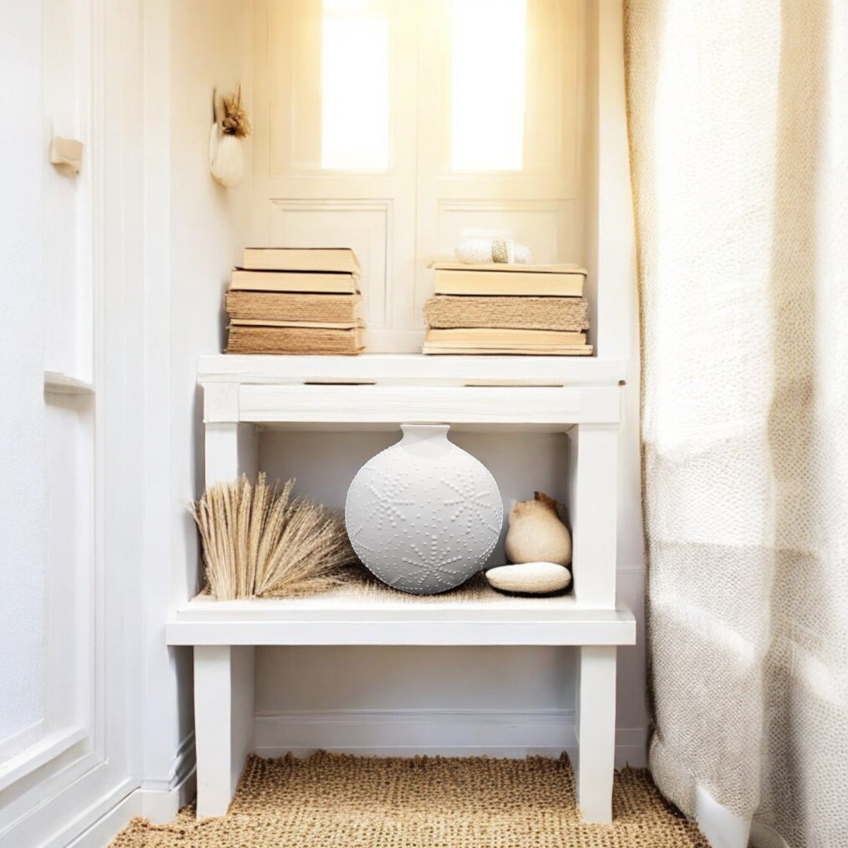 A sunlit white entryway features stacked books on a narrow table with the Sand Dollar Textured Vase in Matte White from Mirabilia Boutique, dried wheat, and a smooth stone on the lower shelf. A woven rug adds coastal charm as sunlight streams in.