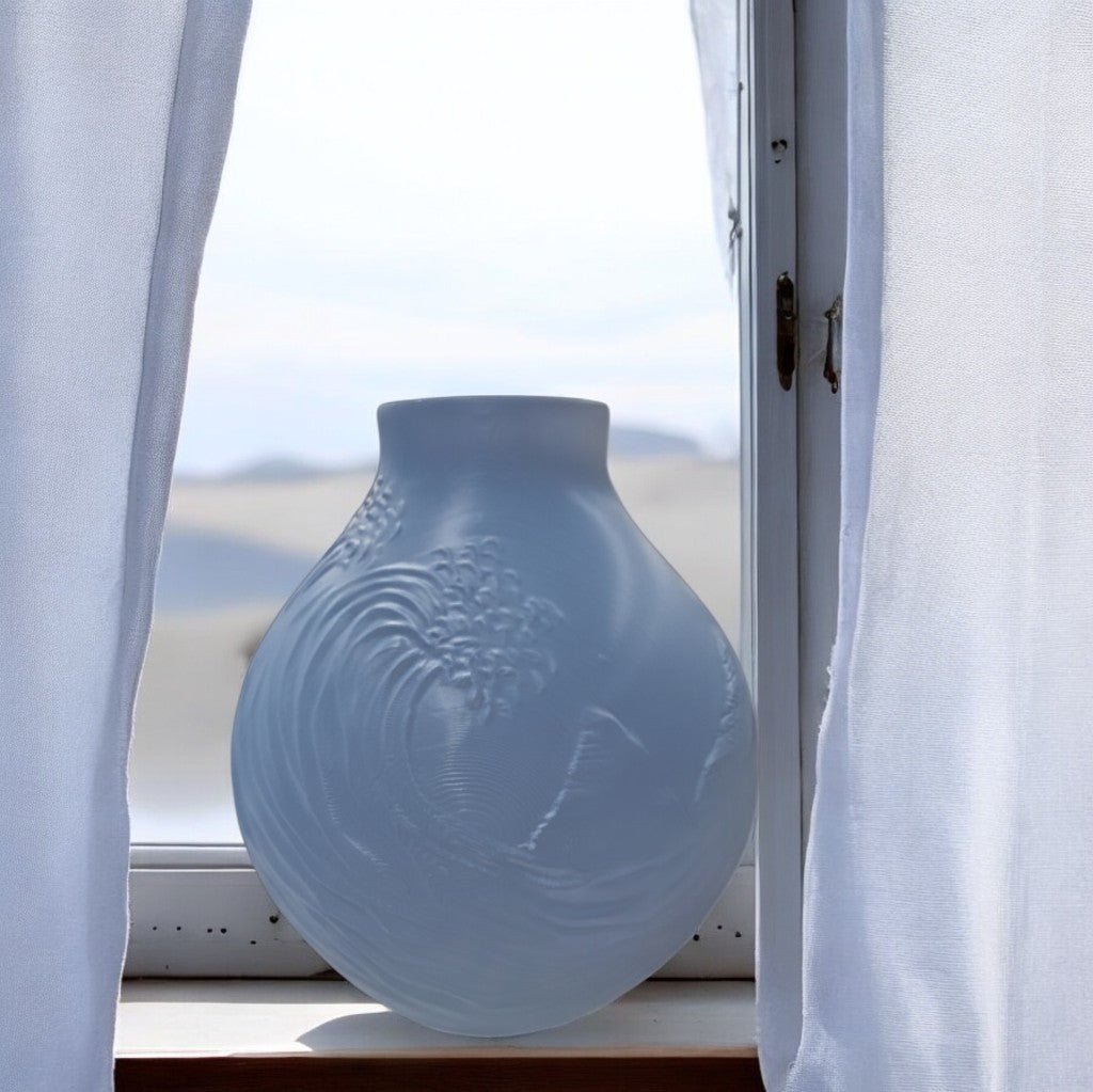 The Stone Blue Coastal Wave Vase by Mirabilia Boutique, featuring delicate engravings and a frosted finish, sits on a windowsill between sheer white curtains with a bright, blurred outdoor view beyond.
