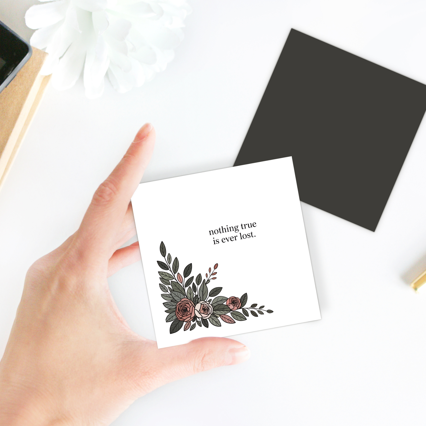 A hand holds the Mirabilia Boutique "Nothing True Is Ever Lost Floral Magnet," featuring floral art and an inspiring message—an ideal encouragement or sympathy gift. Nearby are a black card, a white flower, and desk items.