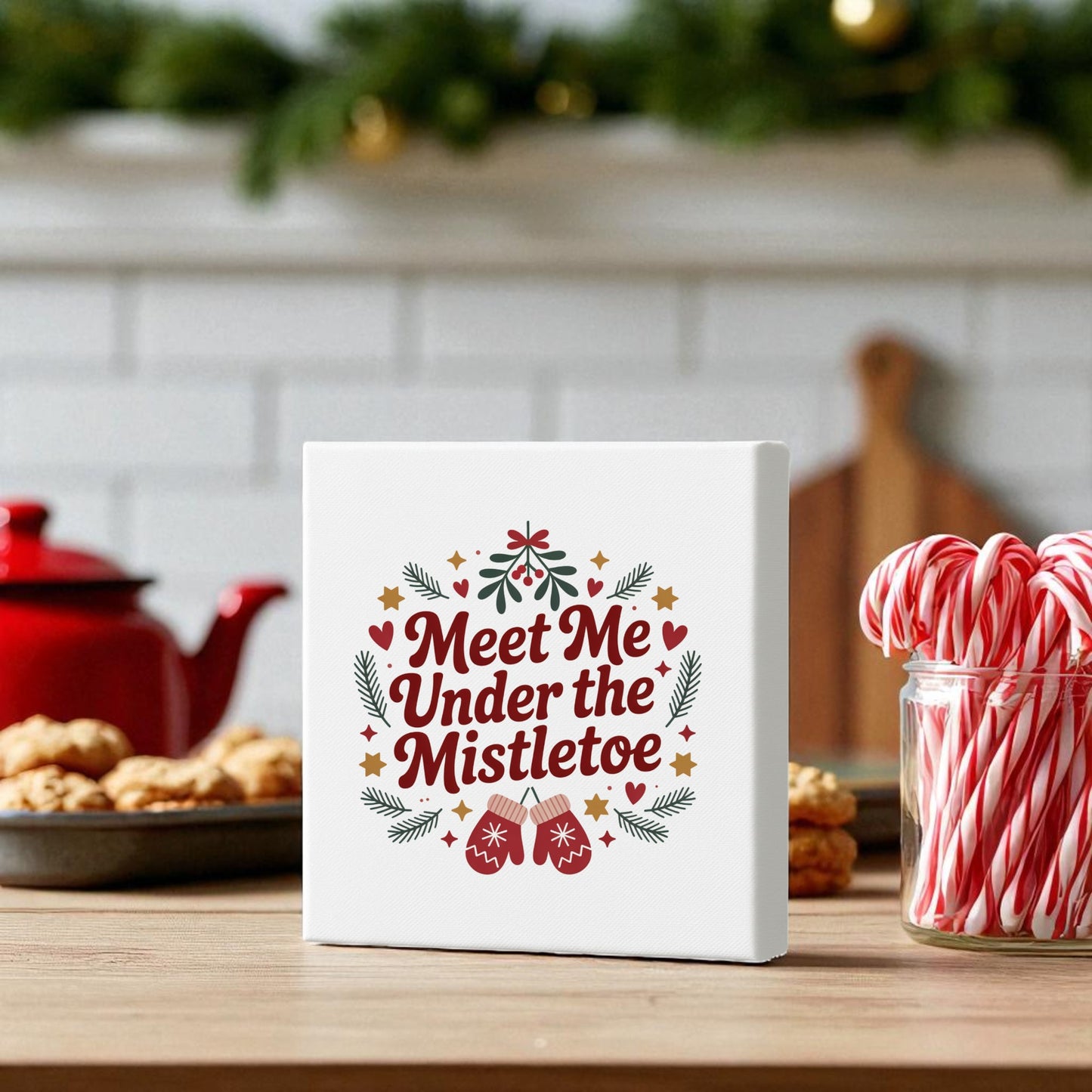 A festive kitchen displays the "Meet Me Under the Mistletoe Canvas Sign" (8x8) from Mirabilia Boutique, surrounded by Christmas decor, cookies, a red teapot, a jar of candy canes, and greenery—ideal holiday gift inspiration.