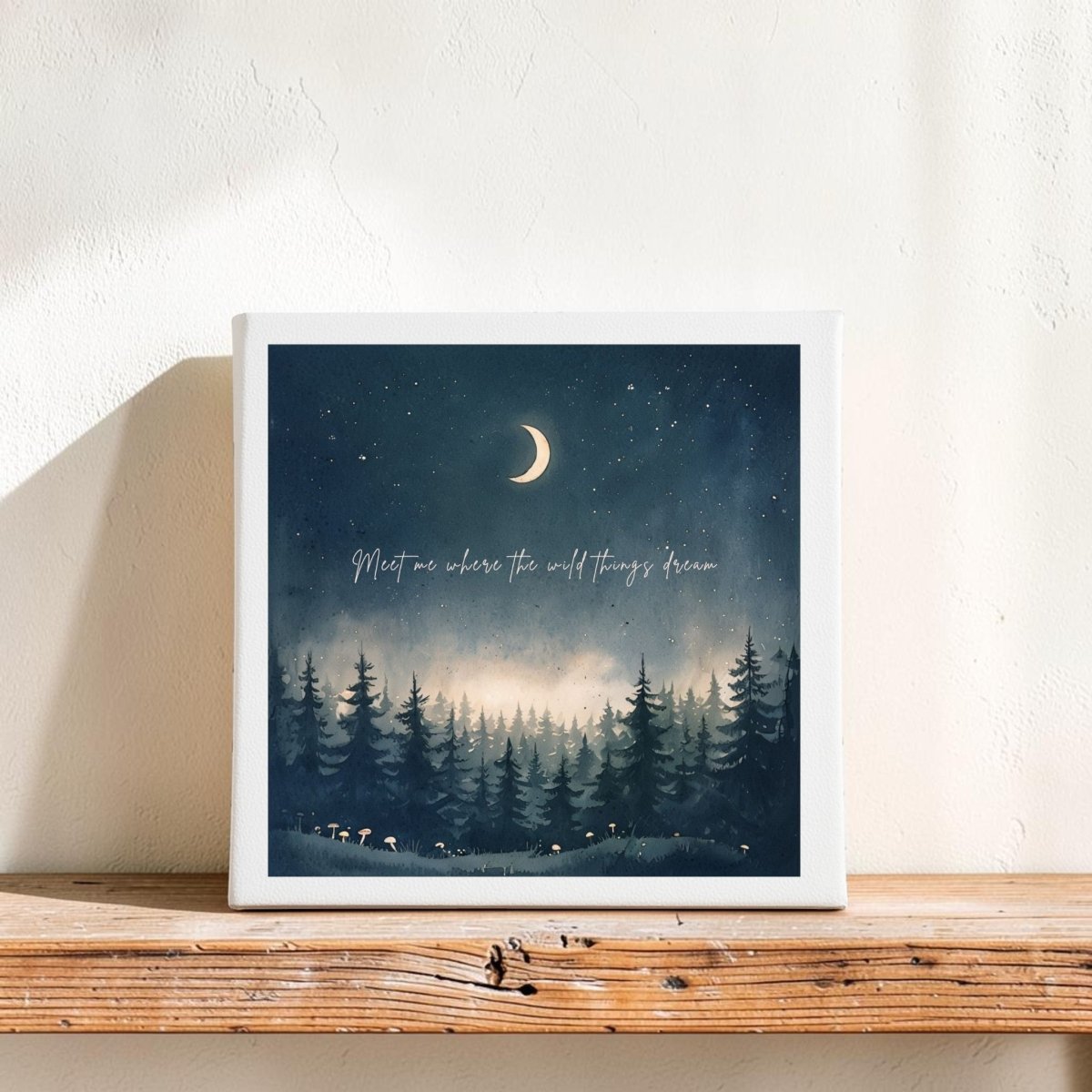 The Meet Me Where the Wild Things Dream Canvas by Mirabilia Boutique features a crescent moon shining in a starry night sky above a pine forest, displayed on a wooden shelf. Across the celestial scene, subtle lettering reads, "Meet me where the wild things dream." This enchanting piece of home decor adds a magical woodland touch to any space.