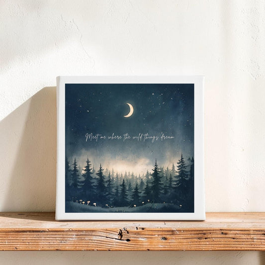 The Meet Me Where the Wild Things Dream Canvas by Mirabilia Boutique features a crescent moon shining in a starry night sky above a pine forest, displayed on a wooden shelf. Across the celestial scene, subtle lettering reads, "Meet me where the wild things dream." This enchanting piece of home decor adds a magical woodland touch to any space.