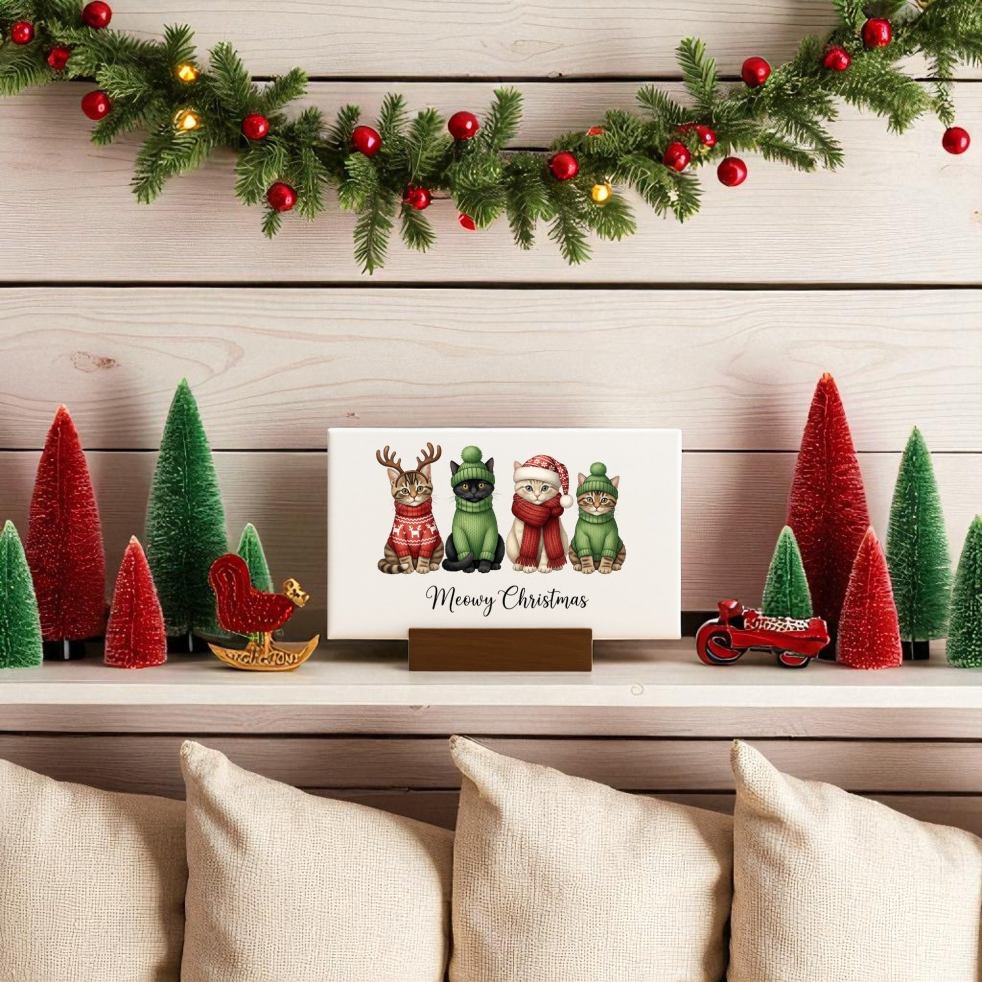 A festive shelf is adorned with red and green Christmas trees, a garland, a wooden squirrel, and a red sled. The Mirabilia Boutique "Meowy Christmas Cat Sign" mini canvas adds cat lover charm beside a sign with three dogs in holiday outfits.